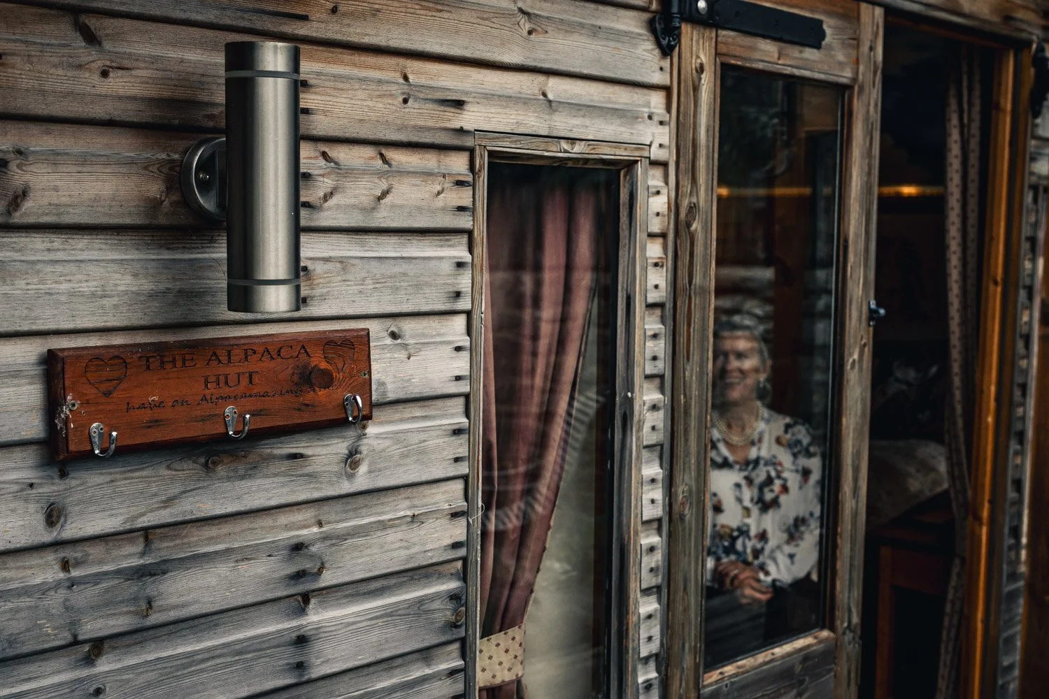 A wooden cabin with a sign that reads 'The Alpaca Hut.' There is a woman inside, visible through a large glass window, smiling and wearing a floral blouse.