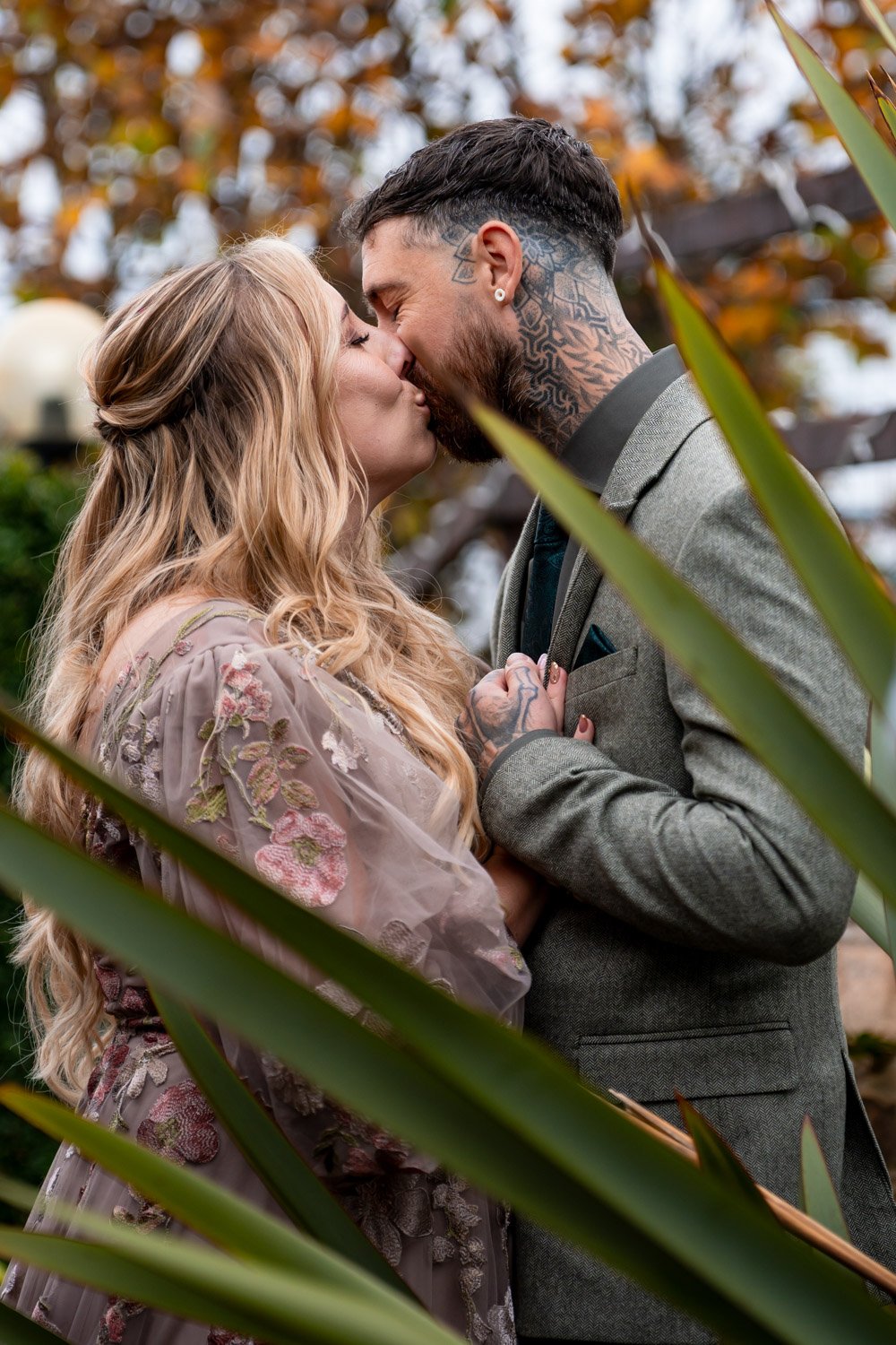 A couple with tattoos kissing outdoors among autumn leaves. The woman has long blonde hair in loose curls and a floral dress. The man has dark hair, a beard, tattoos on his neck and hands, and is wearing a gray suit.
