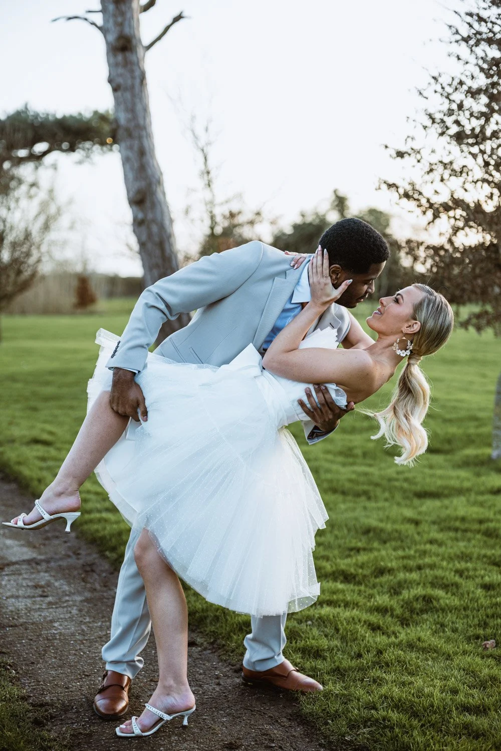 A bride and groom in wedding attire are outdoors on a grassy area. The groom is dipping the bride while they gazing into each other's eyes, holding each other. The background has trees and the sky.