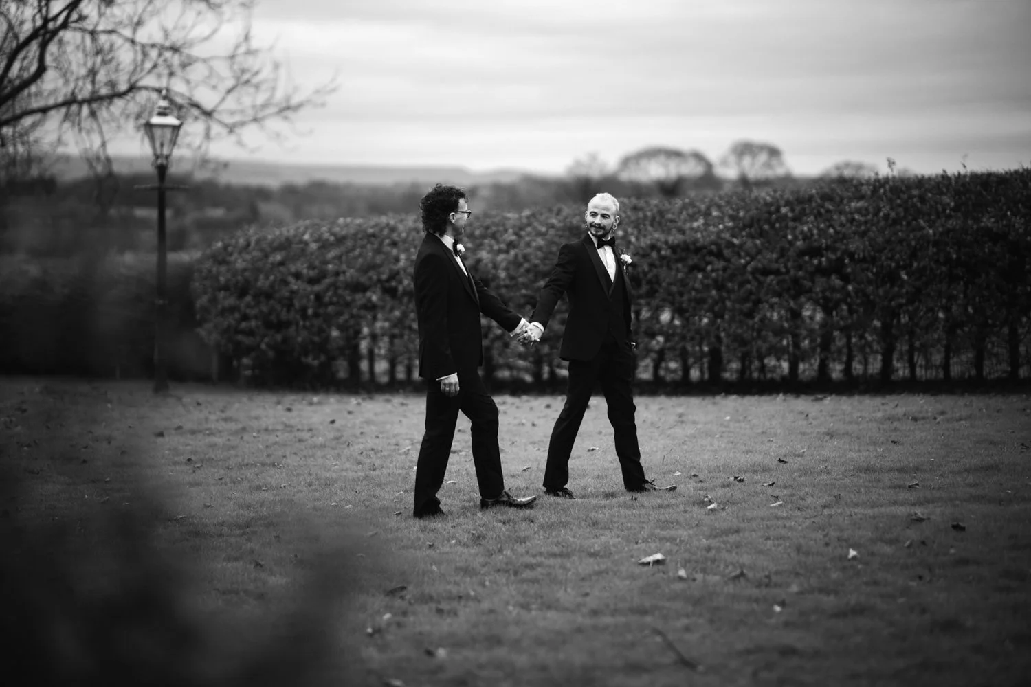 Two men in tuxedos holding hands and walking outdoors in a park or garden with trees and bushes in the background, black and white photo.