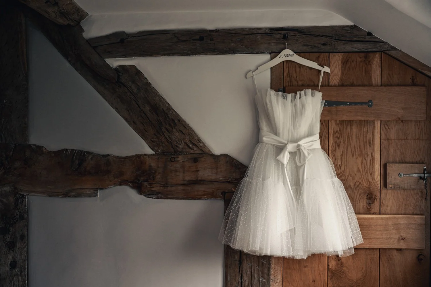 A white wedding dress hanging on a white hanger against a wooden door in a rustic room with exposed wooden beams.