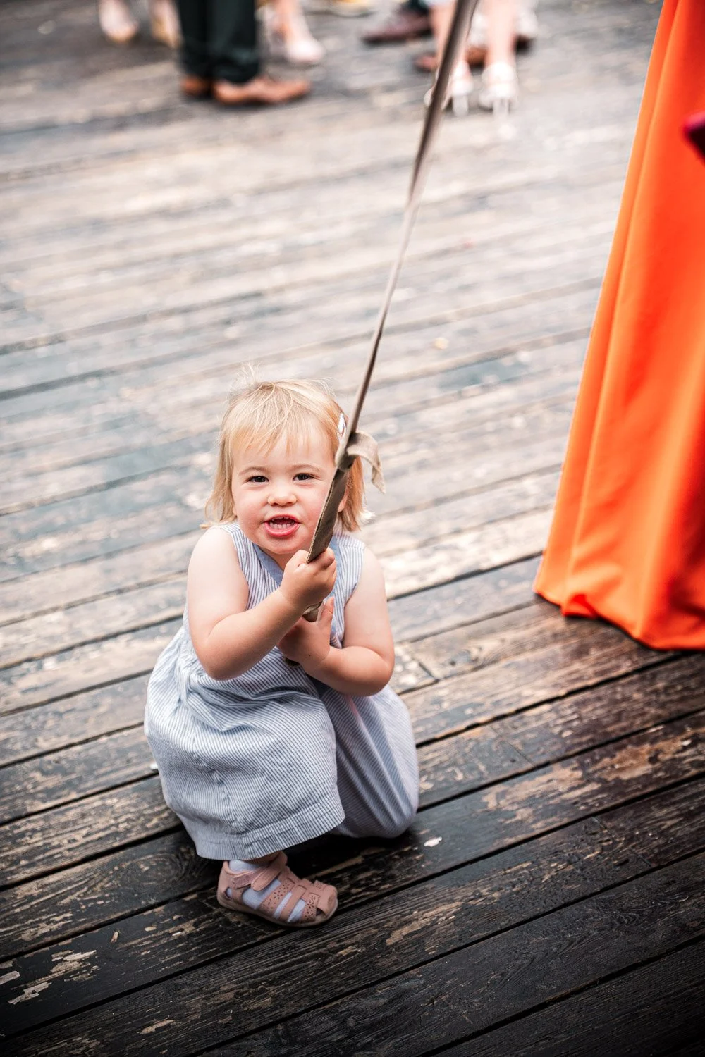A young girl with blonde hair crouches on a wooden deck, holding and pulling on a string attached to an orange fabric or curtain, with other people blurred in the background.