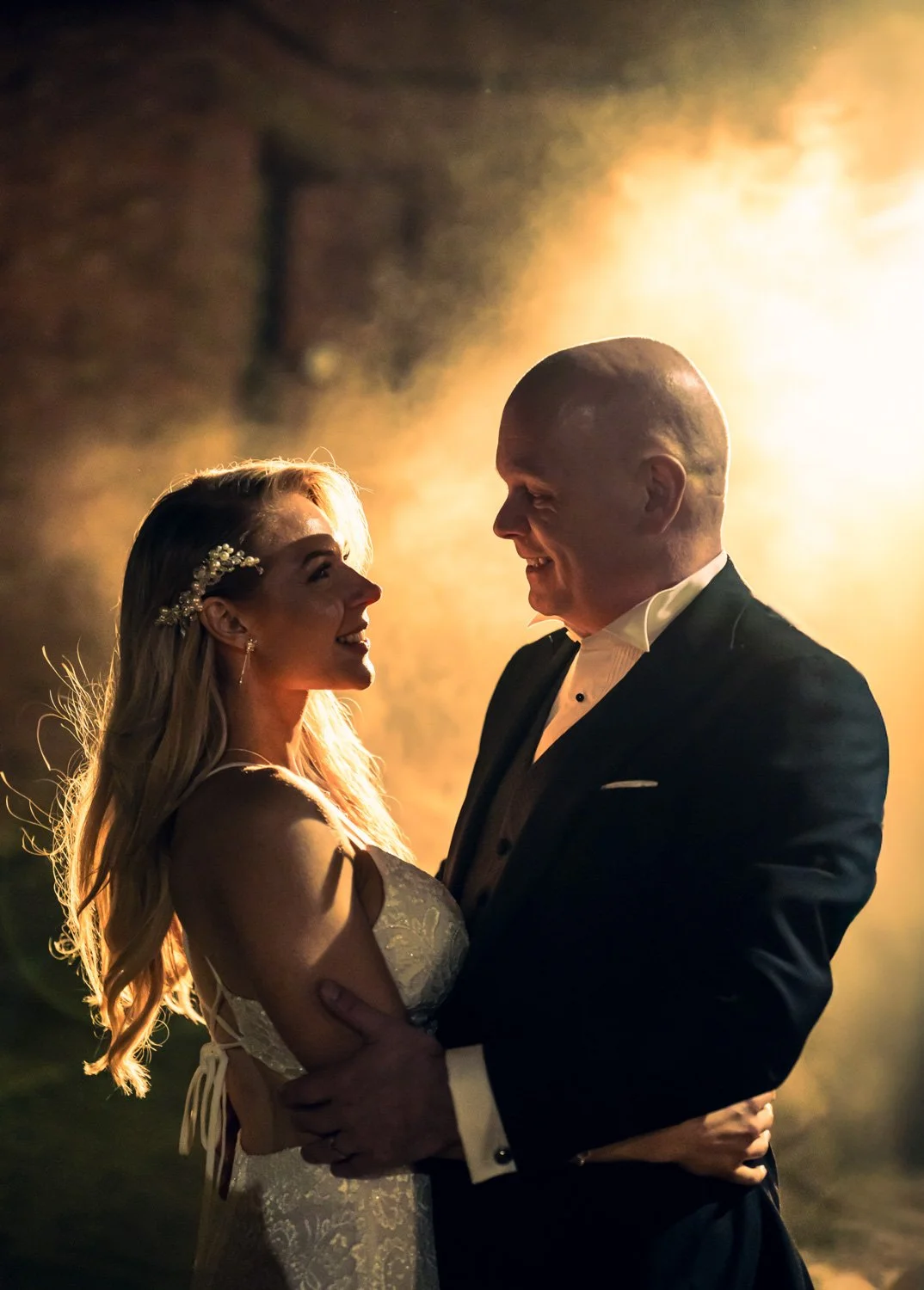 A bride and groom dance at their wedding reception, smiling at each other with warm lighting in the background.