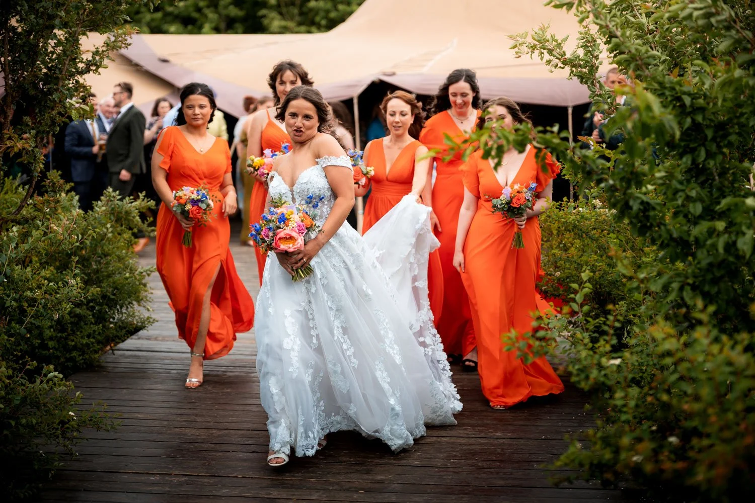 A bride in a white wedding gown holding a colorful bouquet, walking on a wooden pathway with her bridesmaids in matching orange dresses, some holding bouquets, during an outdoor wedding celebration.