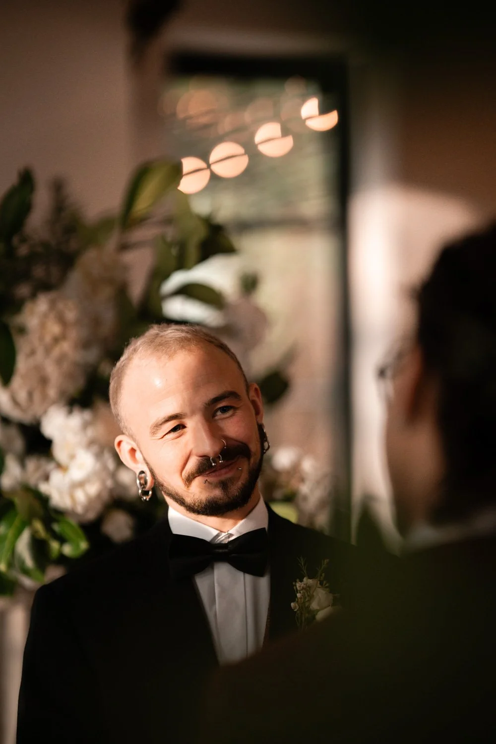 A man in a tuxedo with a bow tie and floral boutonniere smiling during a wedding ceremony, with floral arrangements and blurred background lights.