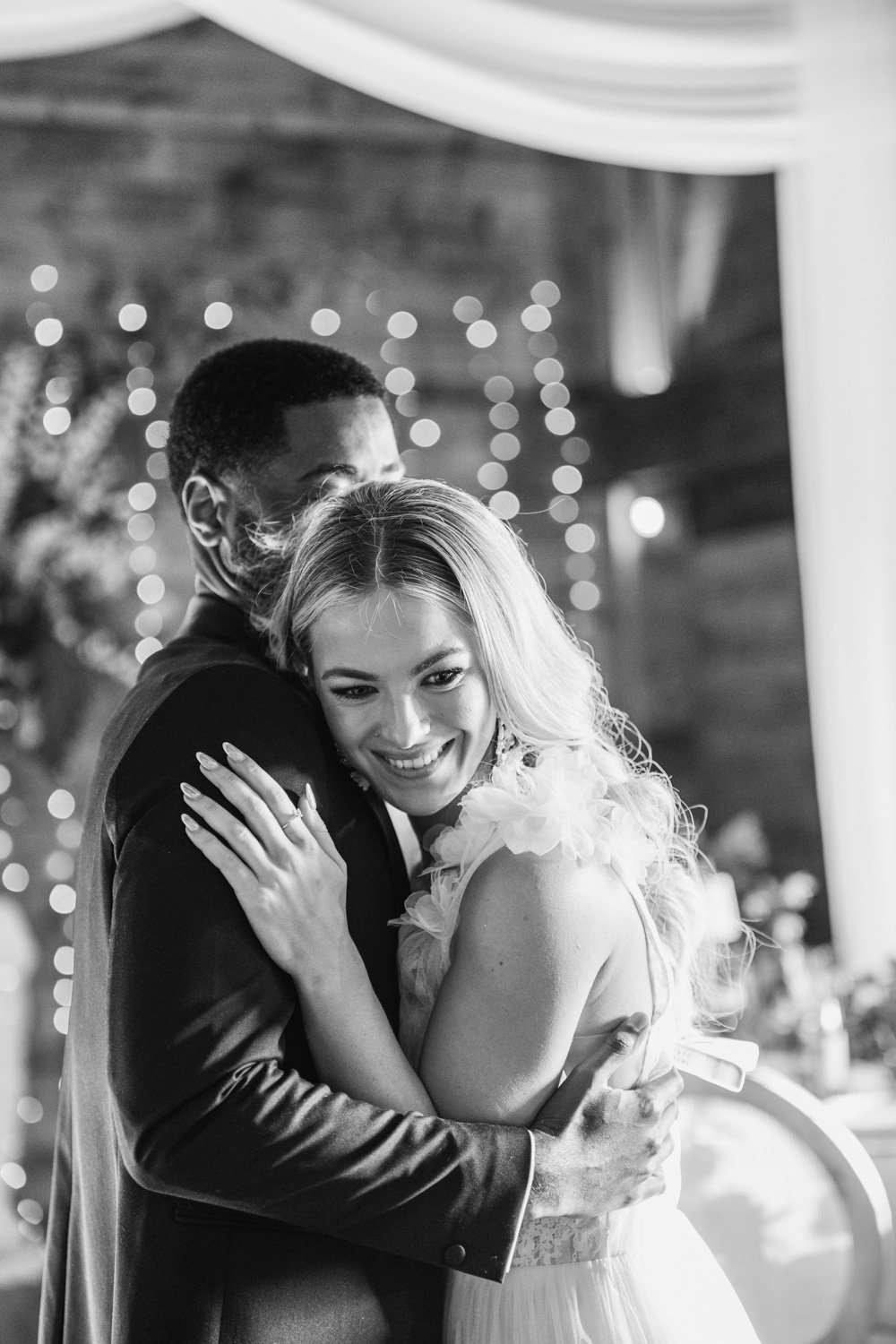 A black and white photo of a man and woman hugging at a wedding celebration, with fairy lights and decor in the background.