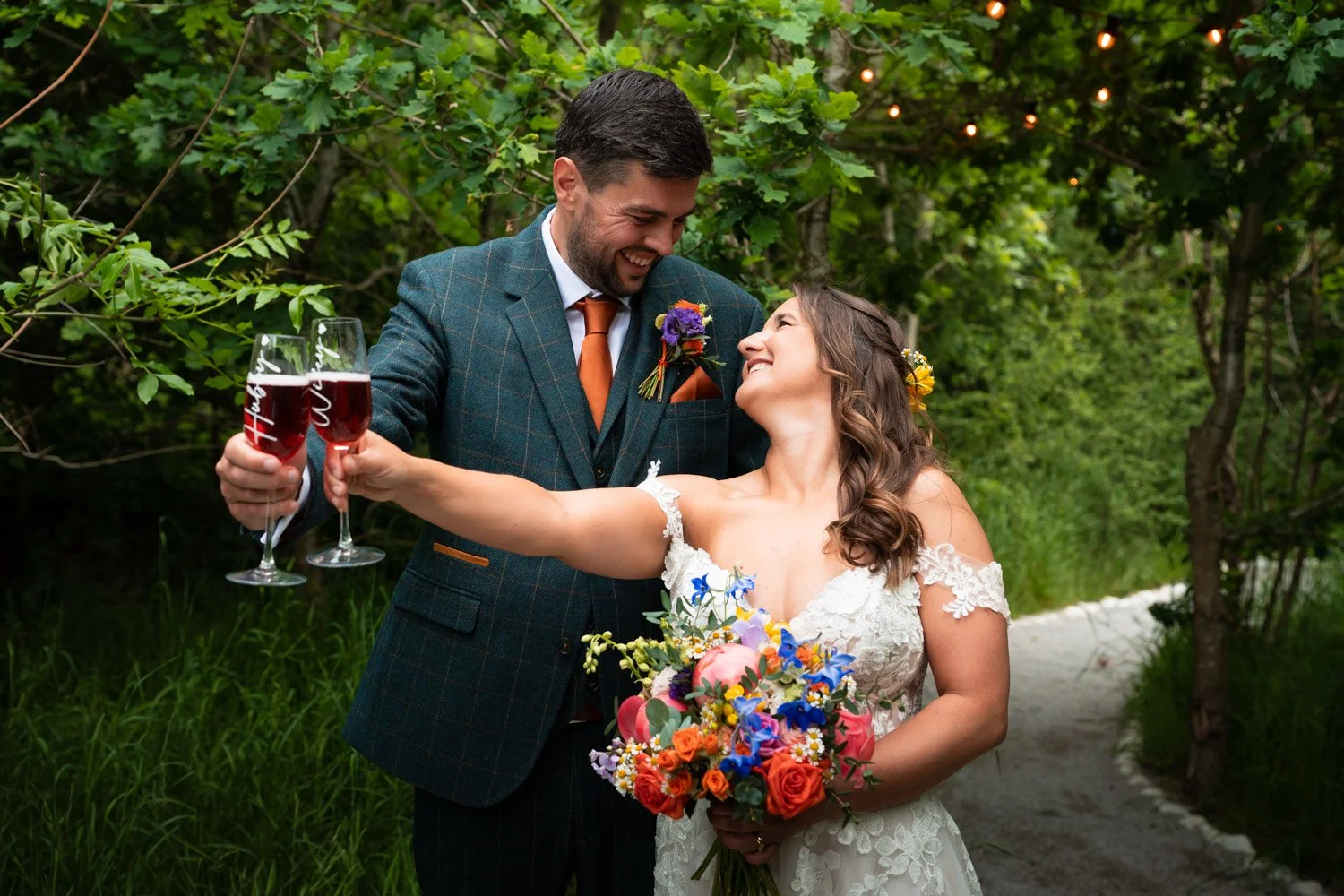 A newlywed couple celebrating outdoors on their wedding day, sharing a joyful moment with the bride holding a bouquet of flowers and the groom holding two glasses of red wine, surrounded by green trees and nature.