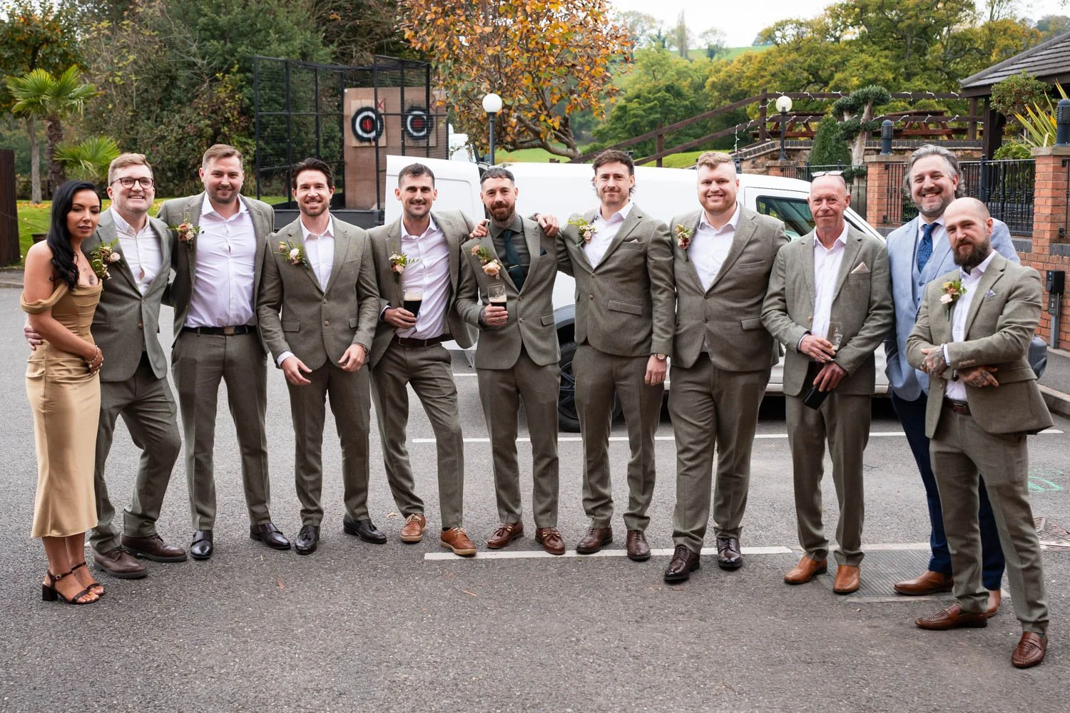A group of people dressed in formal suits and dresses standing together outdoors during daytime, some holding drinks, in front of a white van and decorative background with trees and a brick fence.