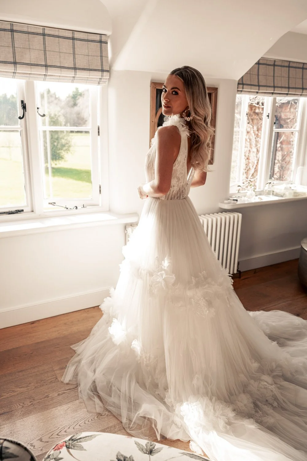 Bridal bride in a white wedding gown standing indoors near windows with sunlight streaming in.