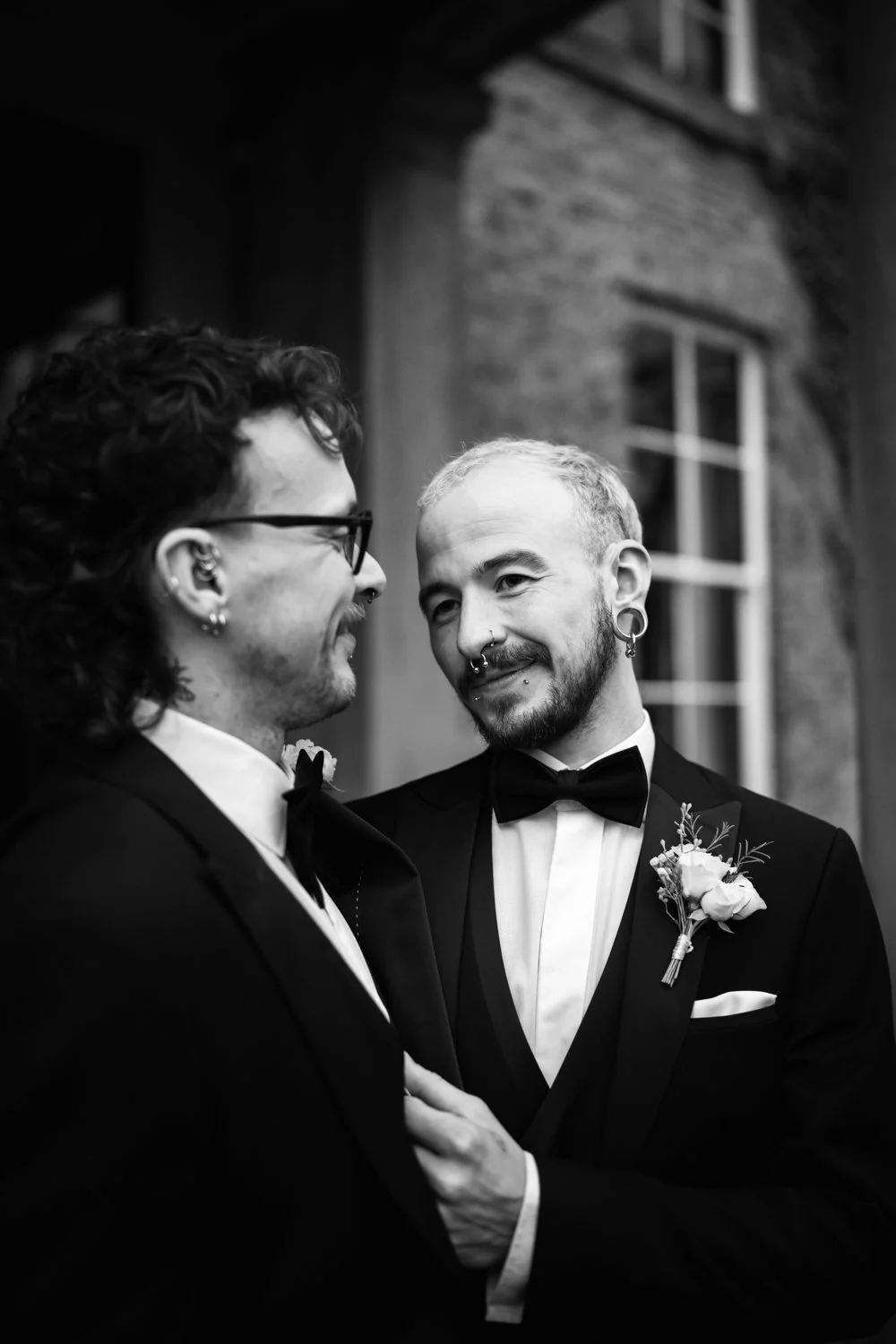 Two men in tuxedos, one with glasses and curly hair, the other with short hair and piercings, sharing a moment at a wedding or formal event, indoors with a brick wall and window in the background.