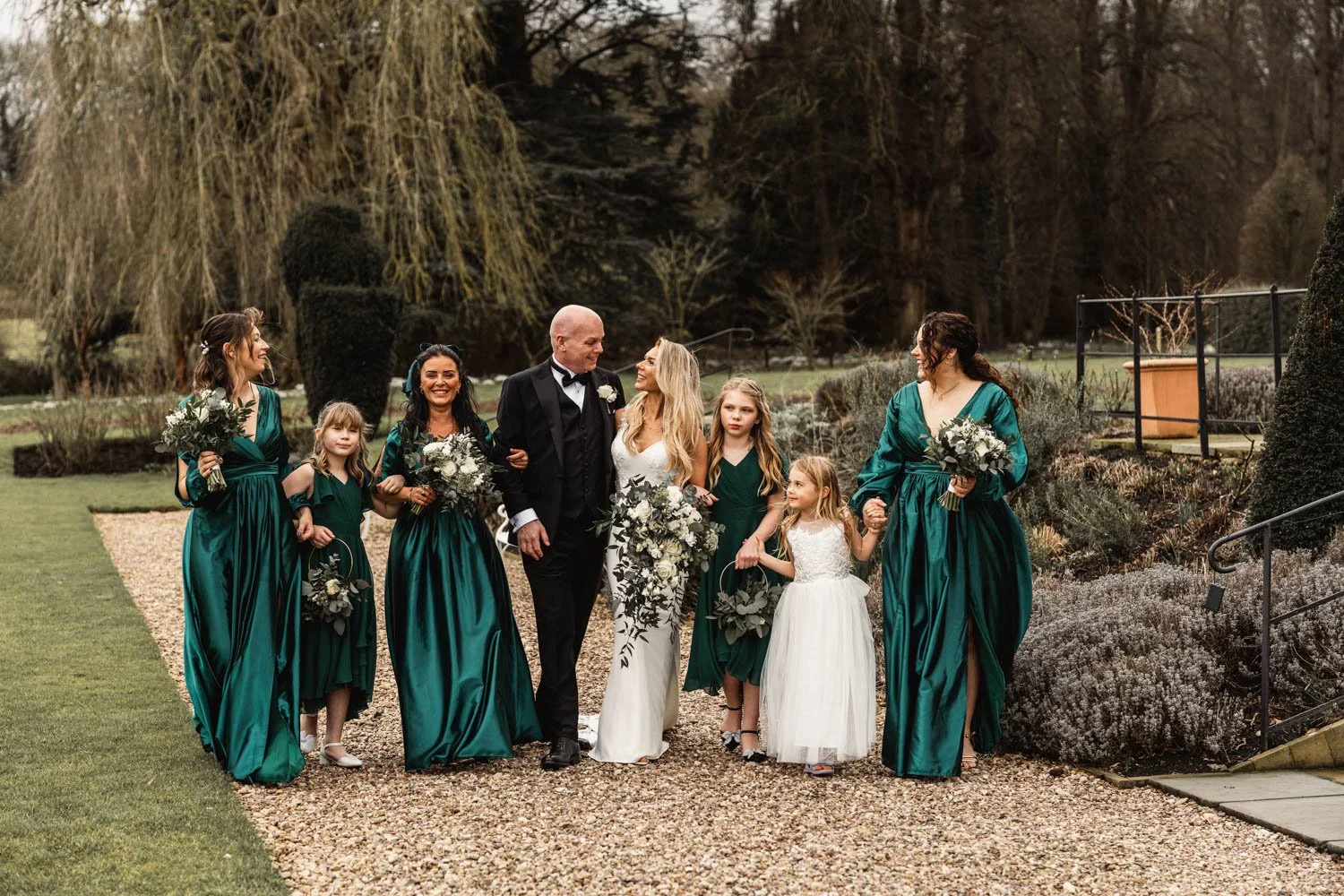 A group of seven women and three young girls walking together outdoors on a wedding day, with the bride and groom in the center, surrounded by family and friends.