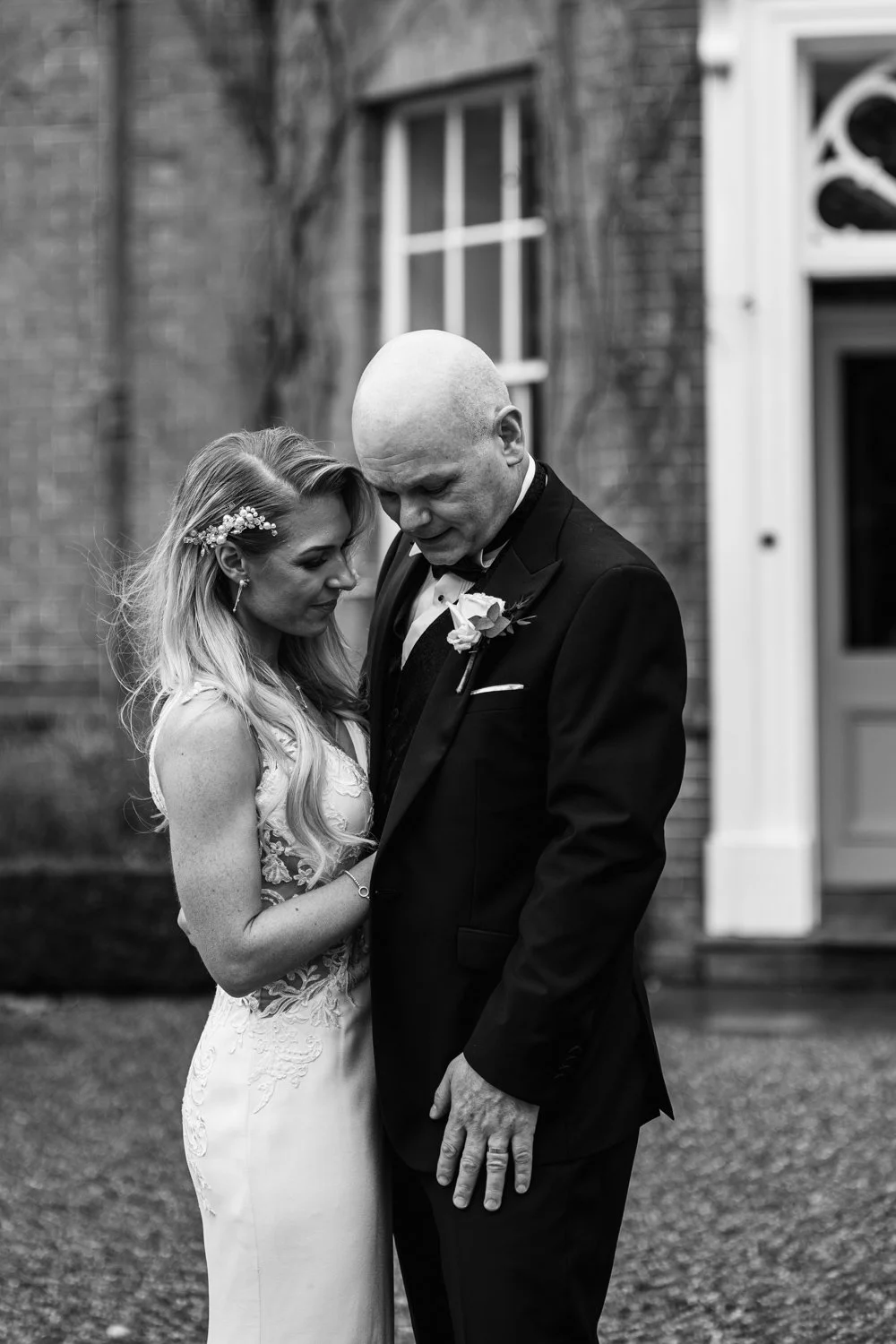 A bride and groom stand close together outdoors, head bowed, in a black-and-white photograph. The bride sports long, styled hair with floral accessories and a lace wedding gown. The groom is dressed in a tuxedo with a boutonniere. They appear emotion
