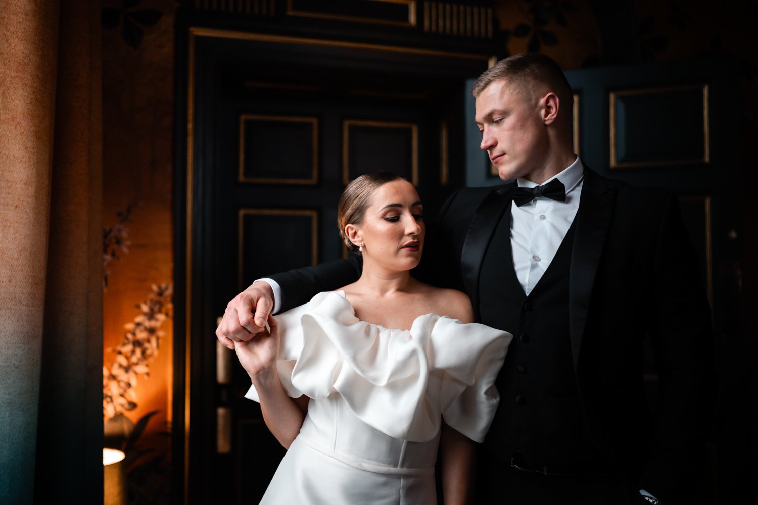 A bride and groom in formal attire, standing close together in a dimly lit, elegant indoor setting with dark walls and gold accents.