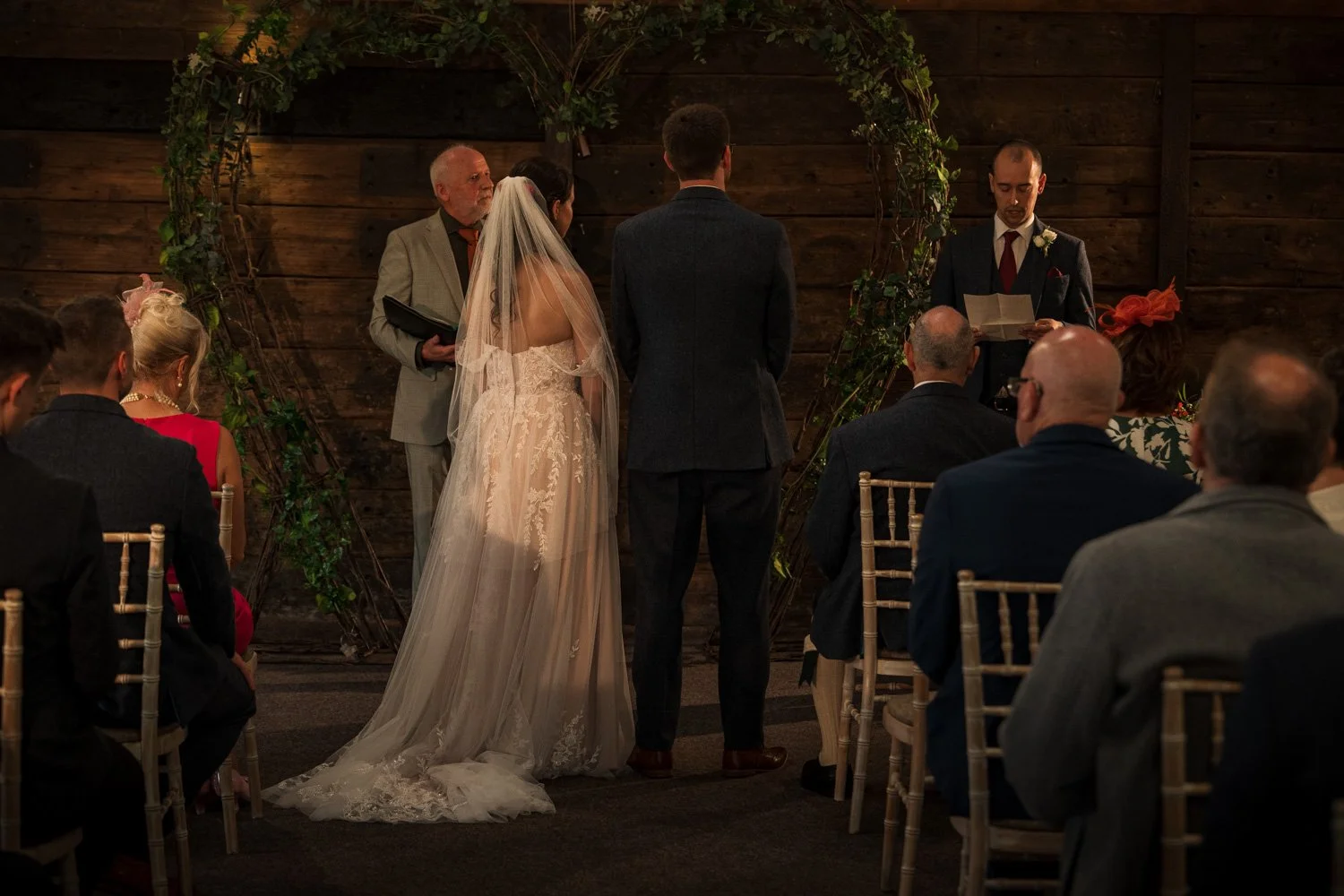 A wedding ceremony with a bride and groom standing before an officiant, surrounded by seated guests in an indoor venue with wooden walls and floral decorations.