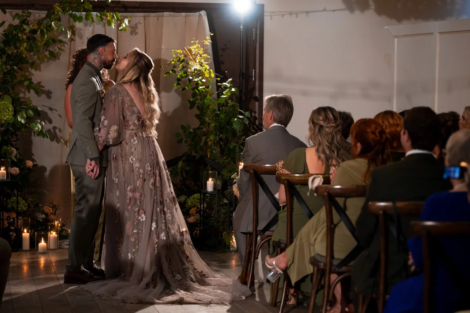 A couple getting married, sharing a kiss during their wedding ceremony in front of seated guests, with candles and green foliage decor.
