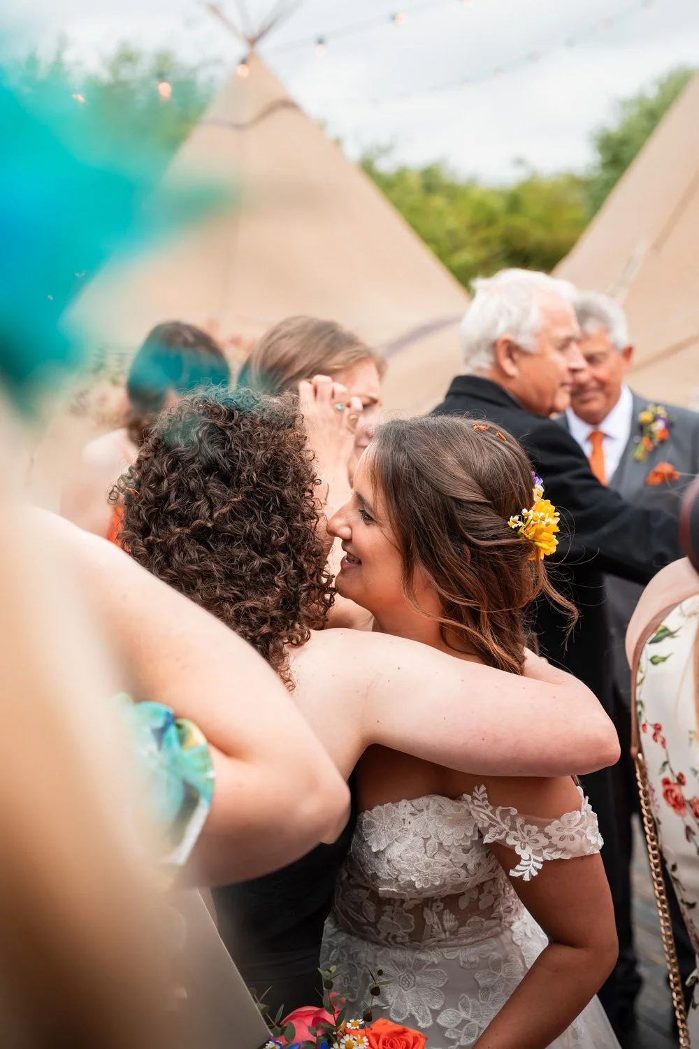 Two women embracing and smiling at each other during a wedding celebration, with people and tents visible in the background.