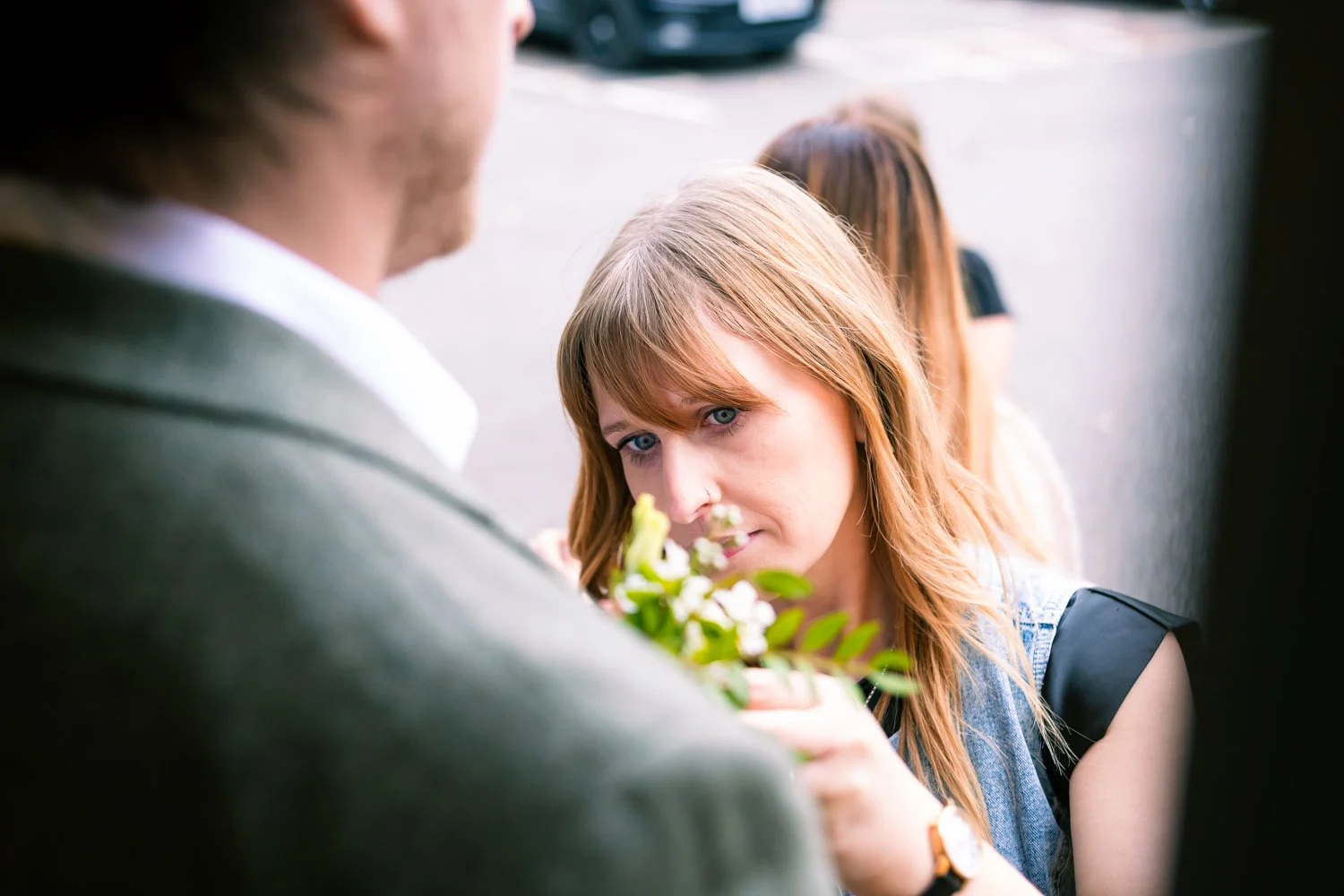 A woman with reddish hair and blue eyes looking at a man holding a small bouquet of white flowers, with other people blurred in the background.