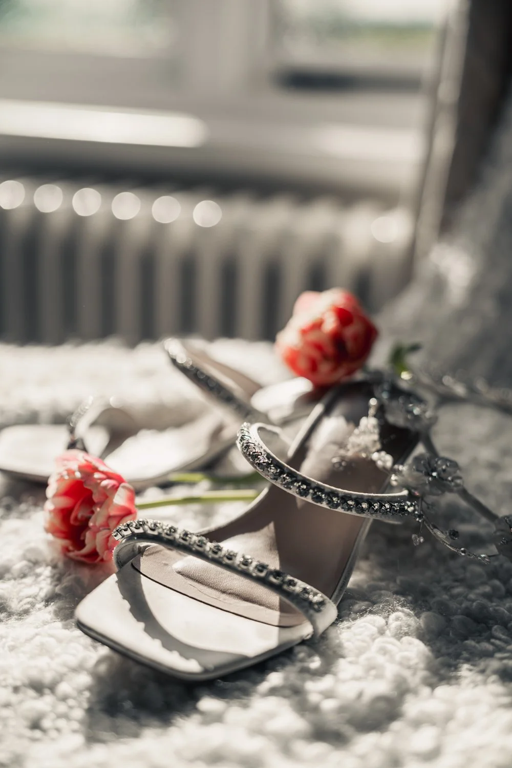 A pair of white high-heeled shoes decorated with rhinestones, with pink and white flowers resting on top, on a textured white surface near a window.