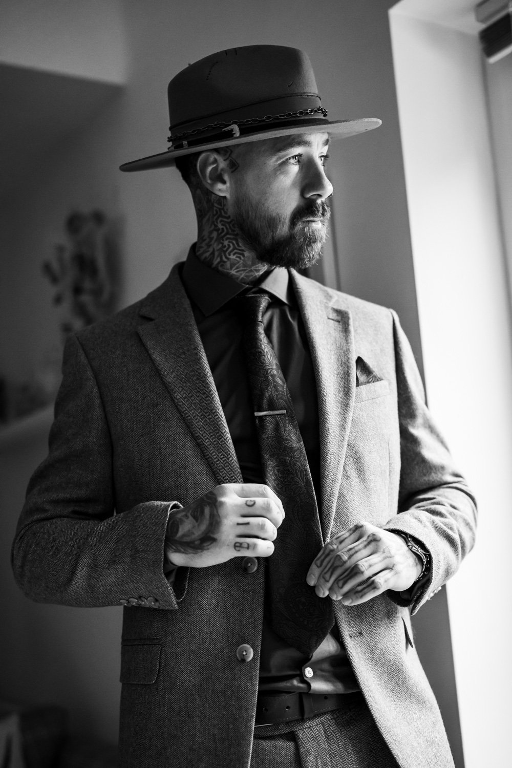 A bearded man with tattoos, dressed in a suit with a tie, wearing a fedora hat, stands near a window looking outside, in black and white.