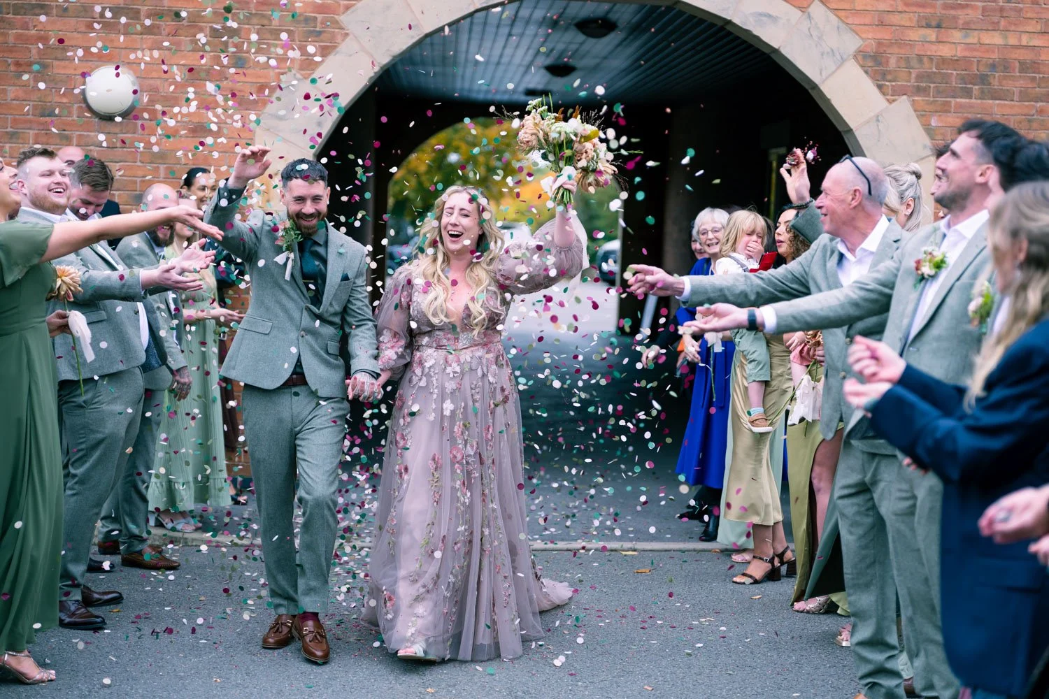 A bride and groom walking through a confetti shower under a brick archway, smiling and holding hands, as friends and family celebrate around them.