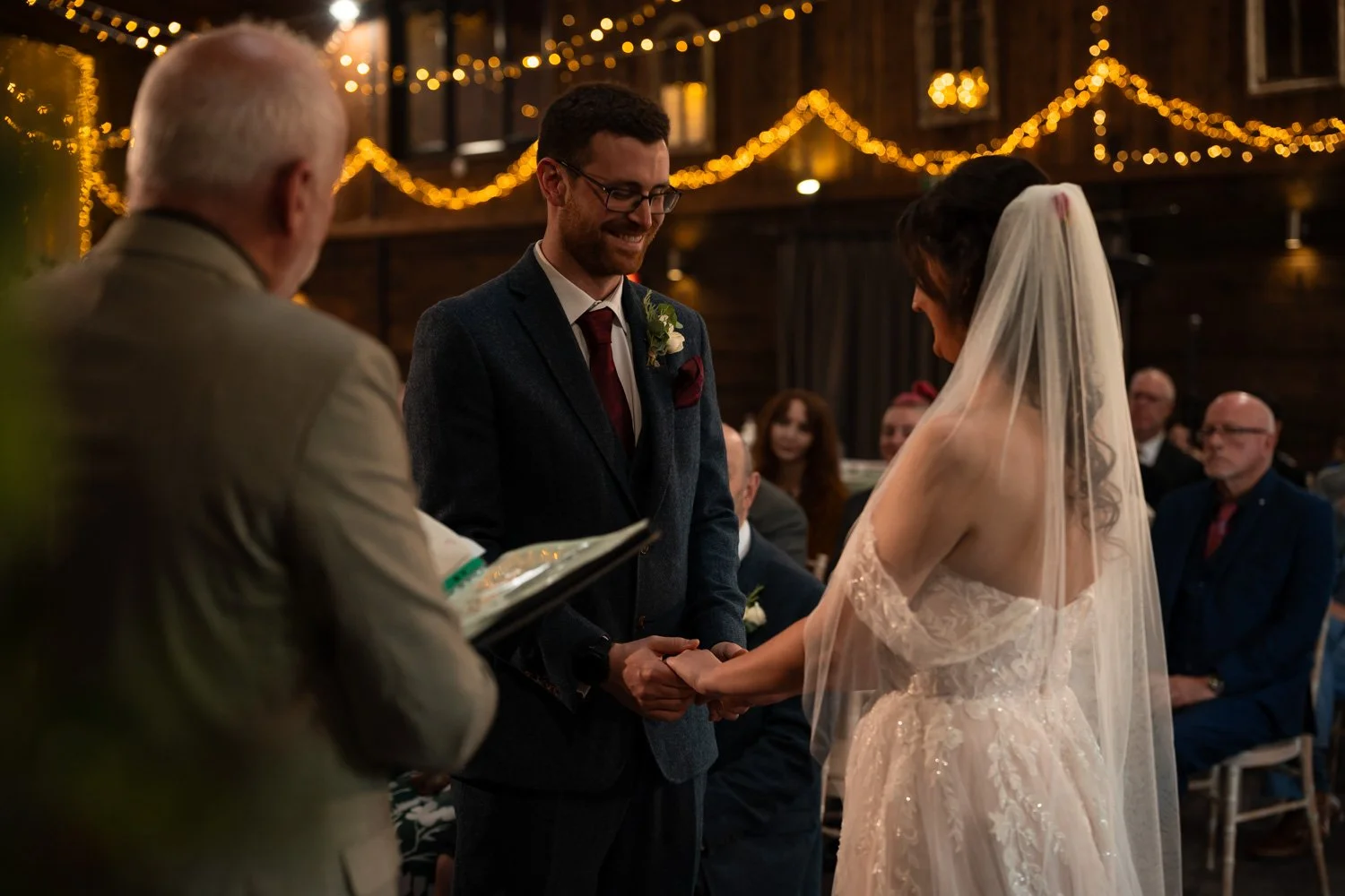 A couple getting married, holding hands, during their wedding ceremony inside a warmly lit venue with string lights, with an officiant present, and guests seated in the background.