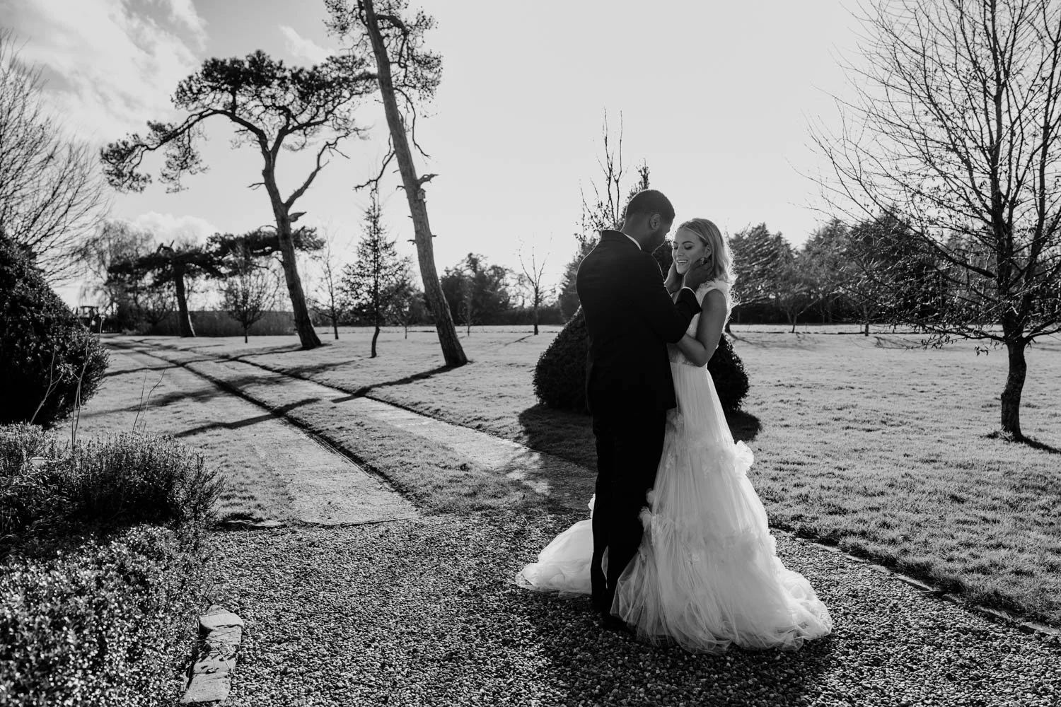 A black and white photo of a bride and groom standing together on a pathway in a park, holding hands and smiling, with trees and grass surrounding them.