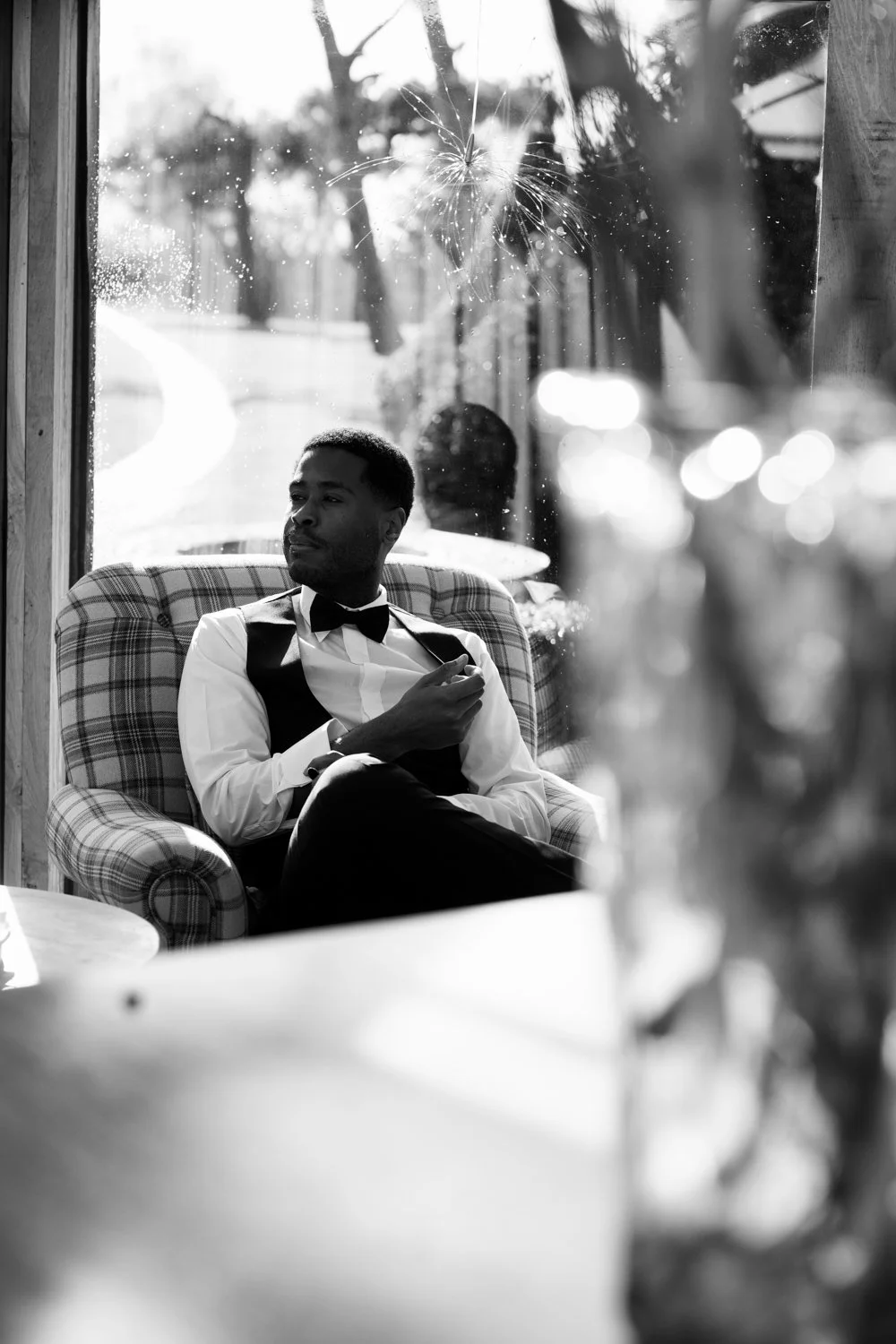 A young man in formal attire sitting in a checked armchair, looking out a window with a broken glass pane, inside a cozy room with blurred outdoor scenery and bokeh lights.