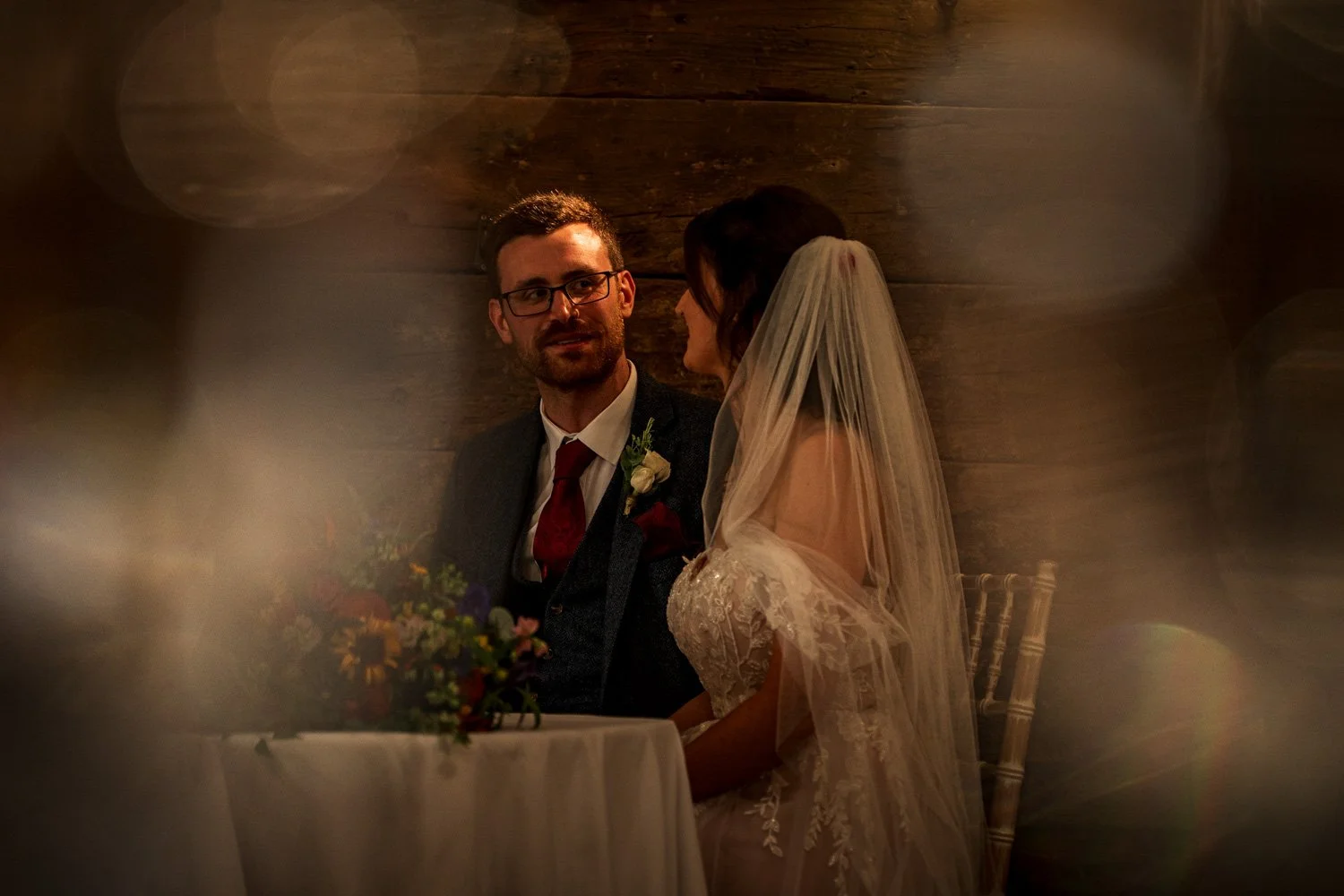 A bride and groom seated at a wedding reception, with the groom wearing glasses, a dark suit, and a red tie, and the bride wearing a lace wedding dress and veil. They are looking at each other against a wooden wall background, with a floral arrangeme