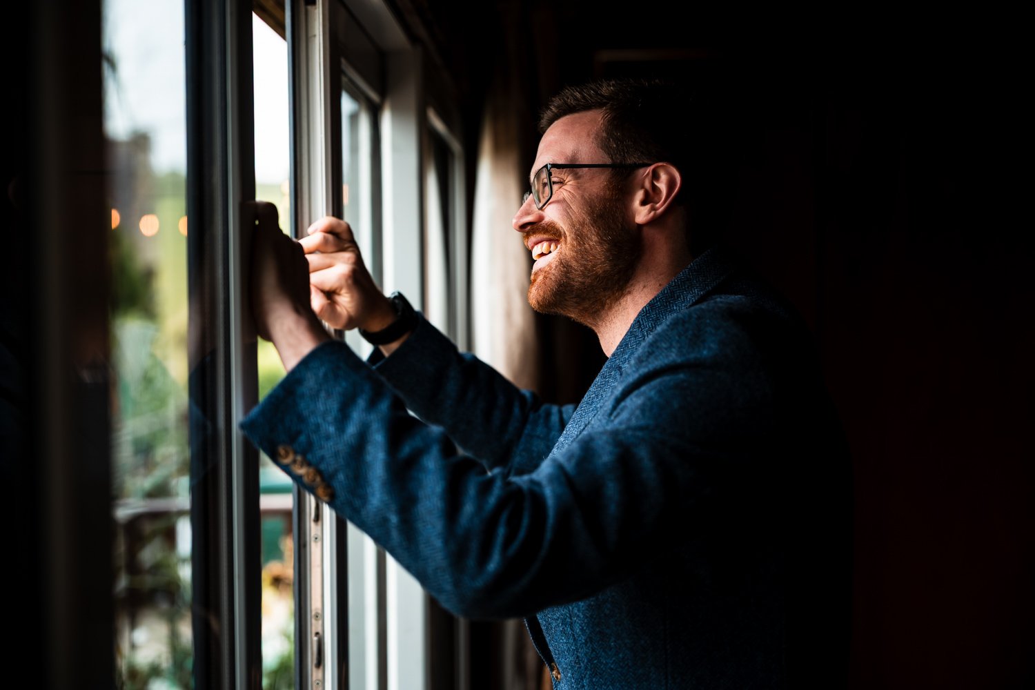 A man wearing glasses and a blue blazer smiling while looking out of a window.