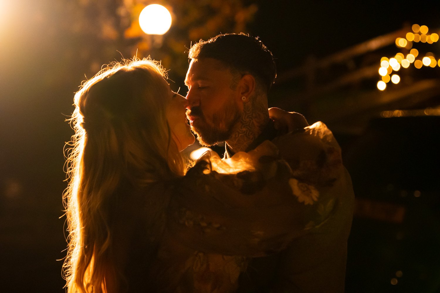 A couple sharing a kiss at night, with warm backlighting and blurred outdoor lights in the background.