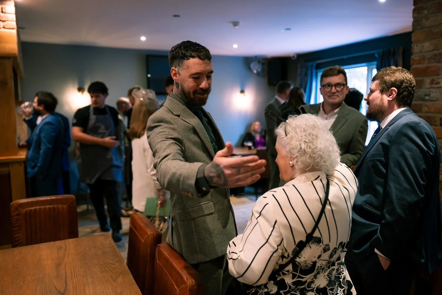 A man in a gray suit shakes hands with an elderly woman with white curly hair, in a crowded indoor space with people socializing in the background.