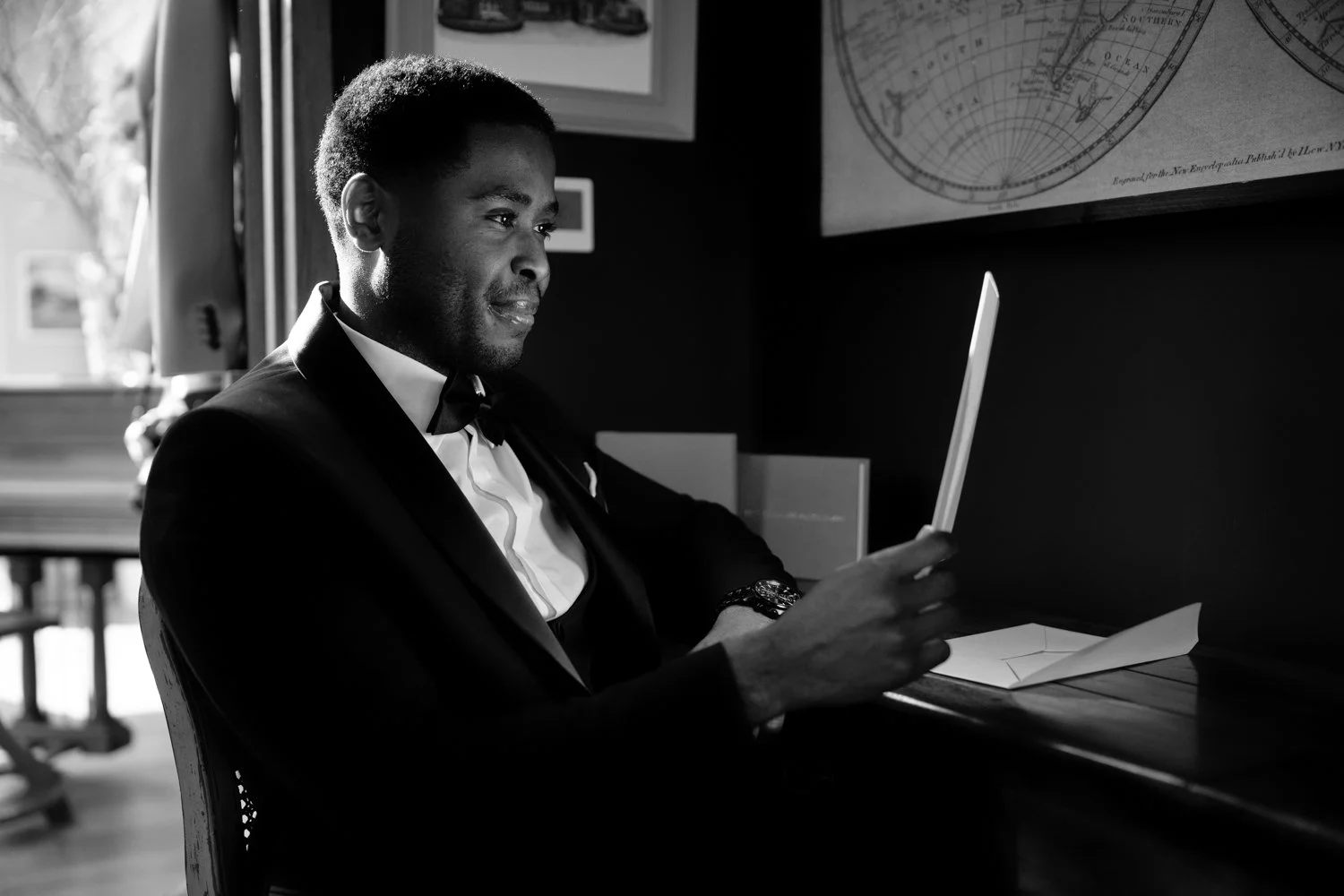 A man in a tuxedo sitting at a desk, looking at a tablet or smartphone, with windows and framed pictures in the background.