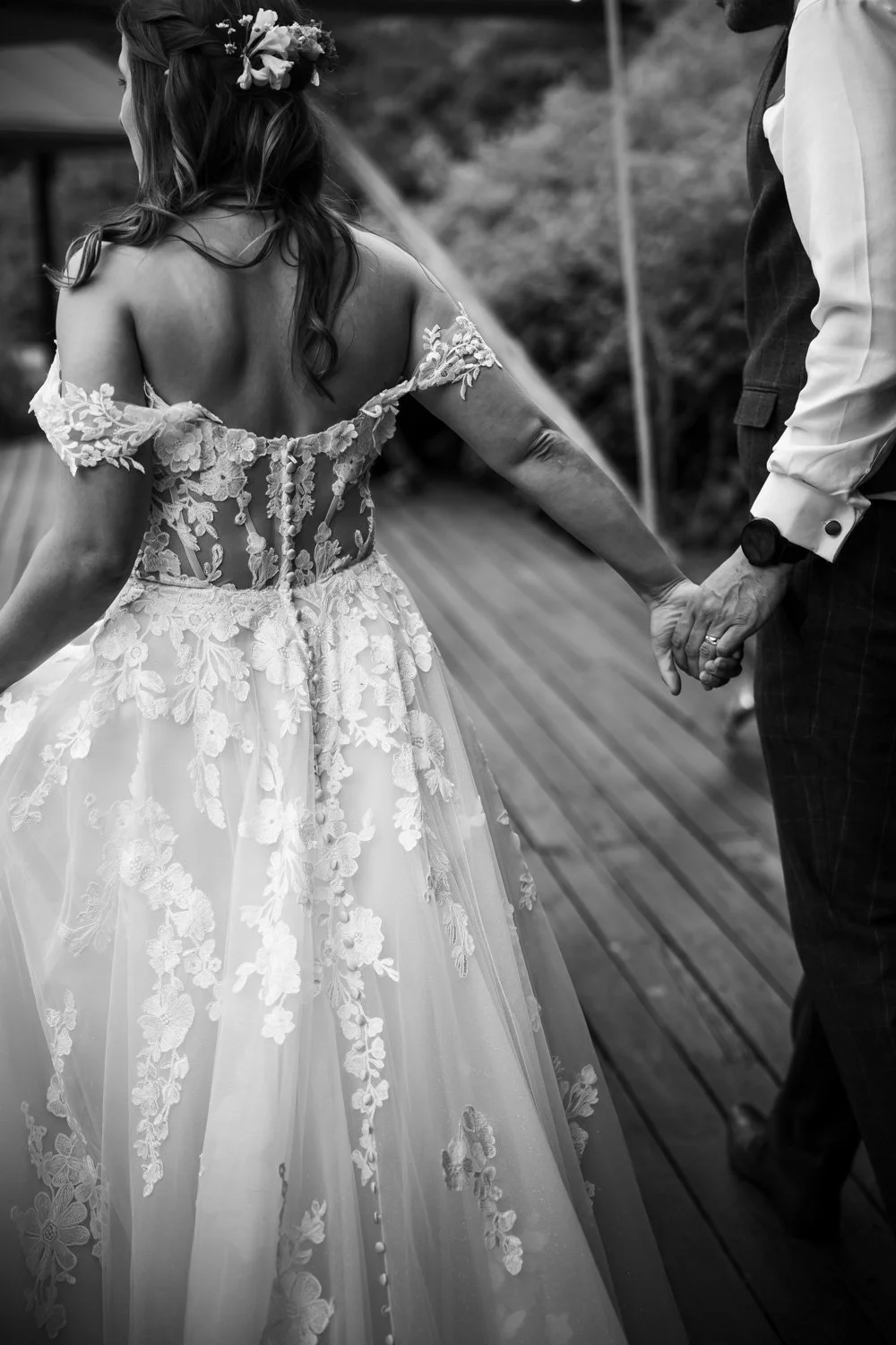 A bride and groom holding hands during their wedding ceremony on a wooden deck. The bride wears a lace-embellished wedding gown with floral details and has a flower crown in her hair. The groom wears a white shirt and dark vest.