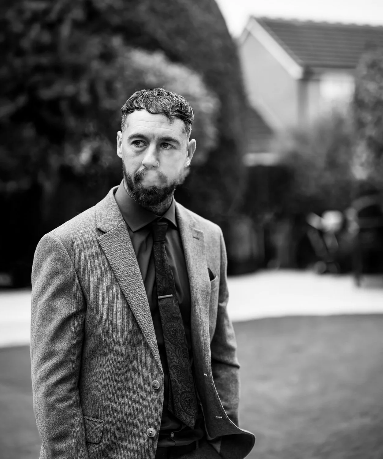 A man with a beard and short hair in a suit smokes a cigarette outdoors on a cloudy day.