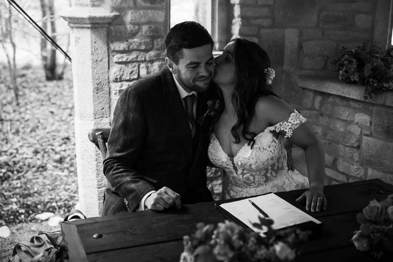 A black and white photo of a bride and groom at their wedding, with the bride kissing the groom on the cheek while sitting at a table with wedding documents.