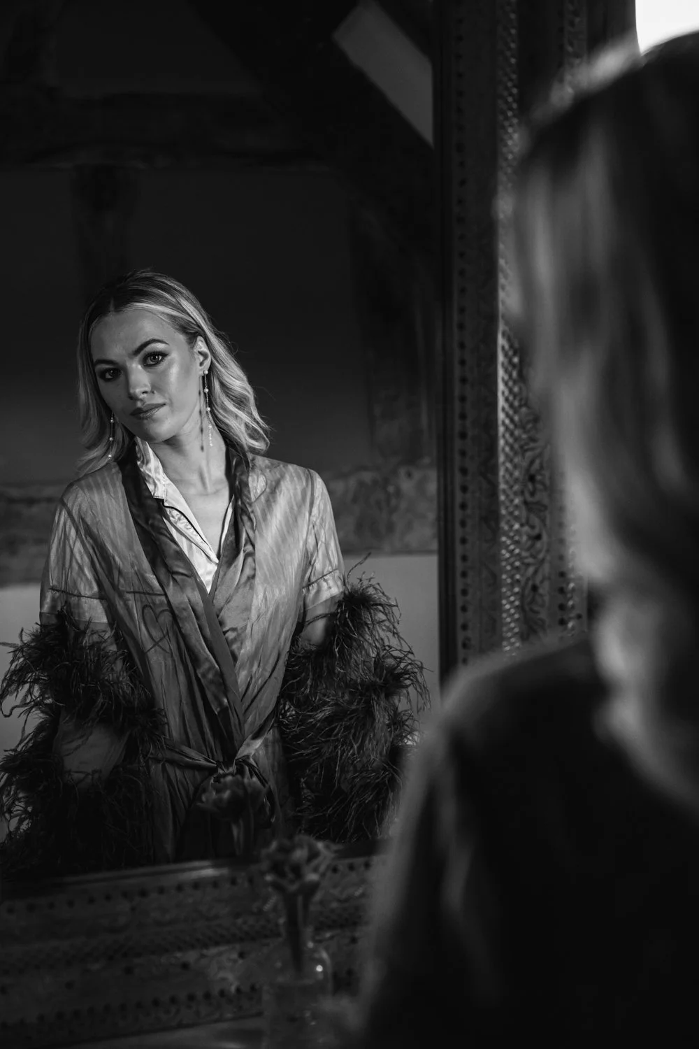 A woman looking at herself in a mirror, wearing a silky robe with feathered sleeves, earrings, and styled hair.