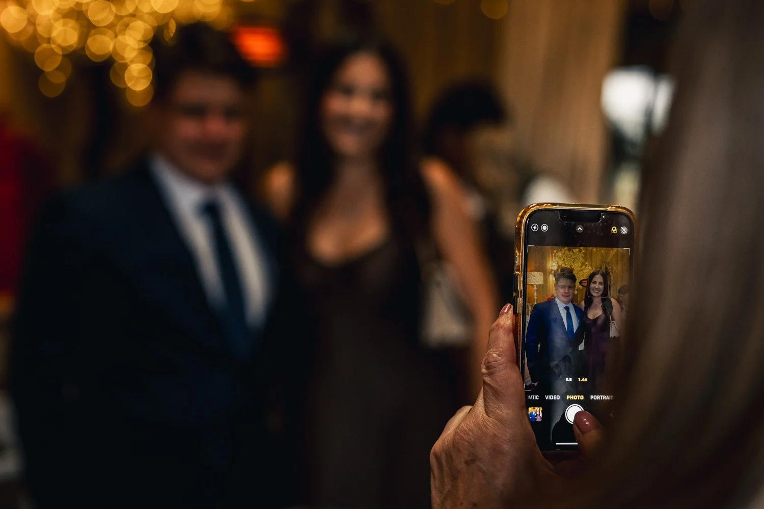 Person taking a photo of two people dressed in formal attire at an indoor event, with a blurred background of warm lights.