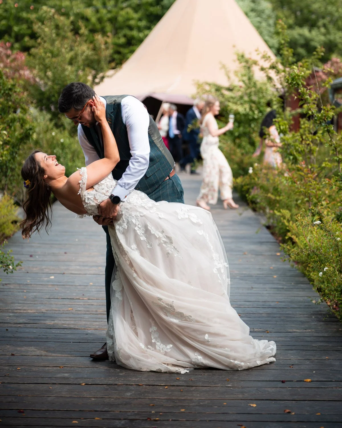 A joyful bride and groom dancing on a wooden pathway outdoors at a wedding celebration. The bride is wearing a lace wedding gown and the groom dressed in a checked vest and glasses. In the background, guests are socializing near greenery and a tent.