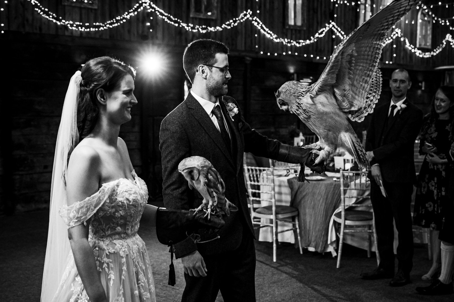 A wedding ceremony with a man and woman, possibly a bride and groom, holding trained owls on their gloved hands, with guests observing in the background. Black and white photo, indoor rustic setting with string lights.