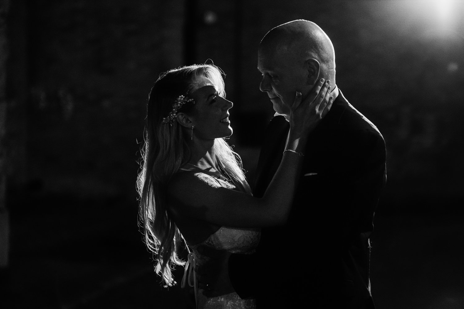 Black and white photo of a bride and groom dancing, with the bride touching the groom’s face, smiling, and the groom looking at her.