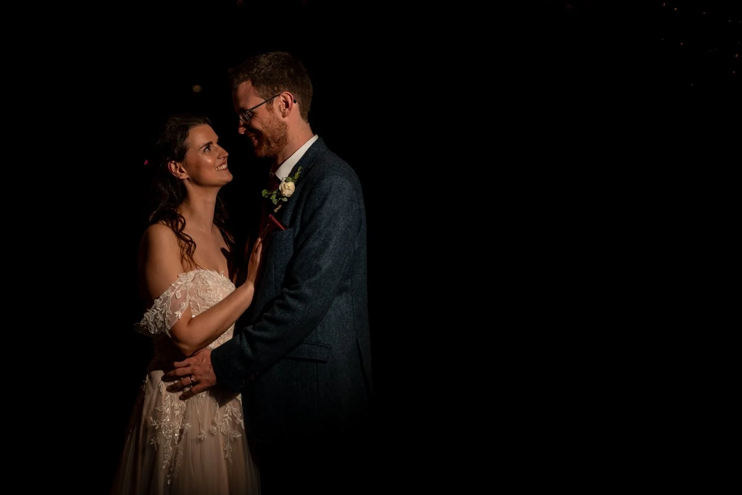 A bride and groom on their wedding day standing close, smiling and gazing into each other's eyes against a black background.