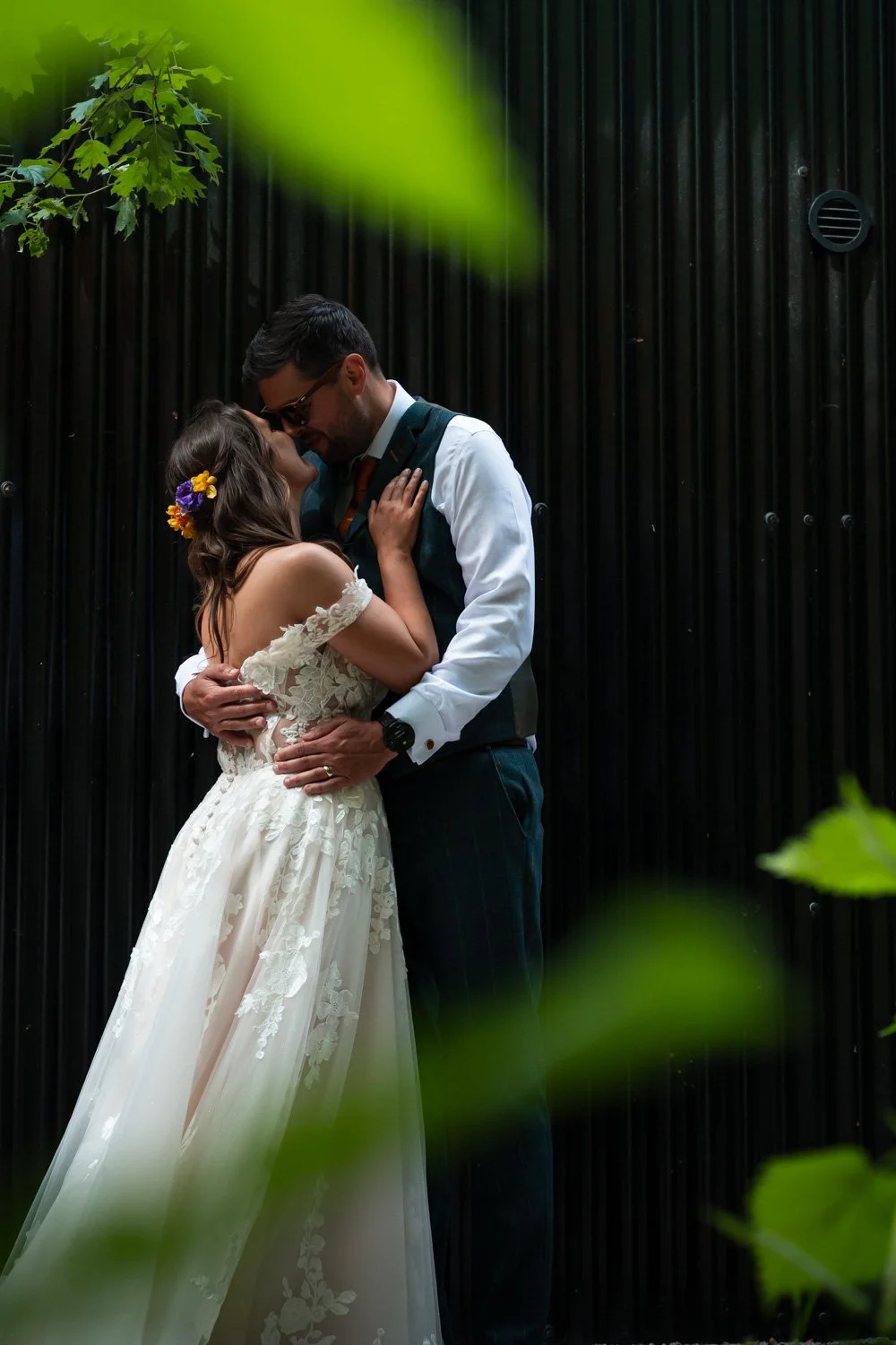 A couple embraces in wedding attire against a black corrugated wall, with green leaves in the foreground.