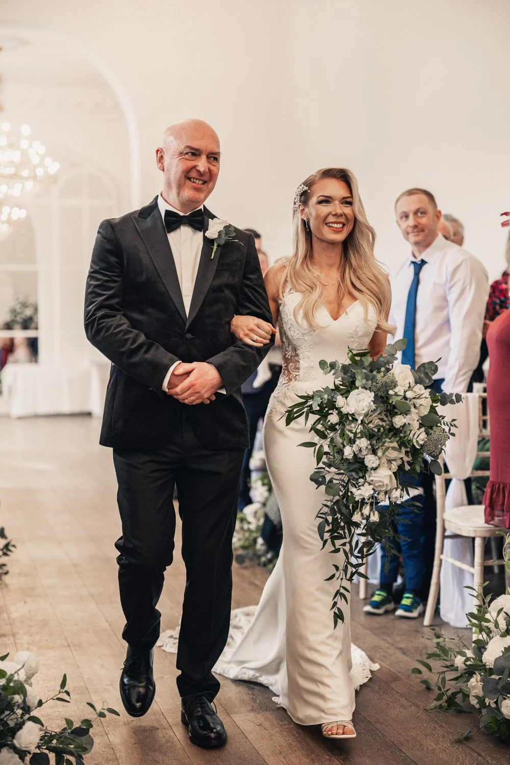 A bride with long blonde hair and a floral hairpiece, smiling and holding a large bouquet of white flowers, walking down the aisle with a groom at her side, during a wedding ceremony in a well-lit, elegant venue.