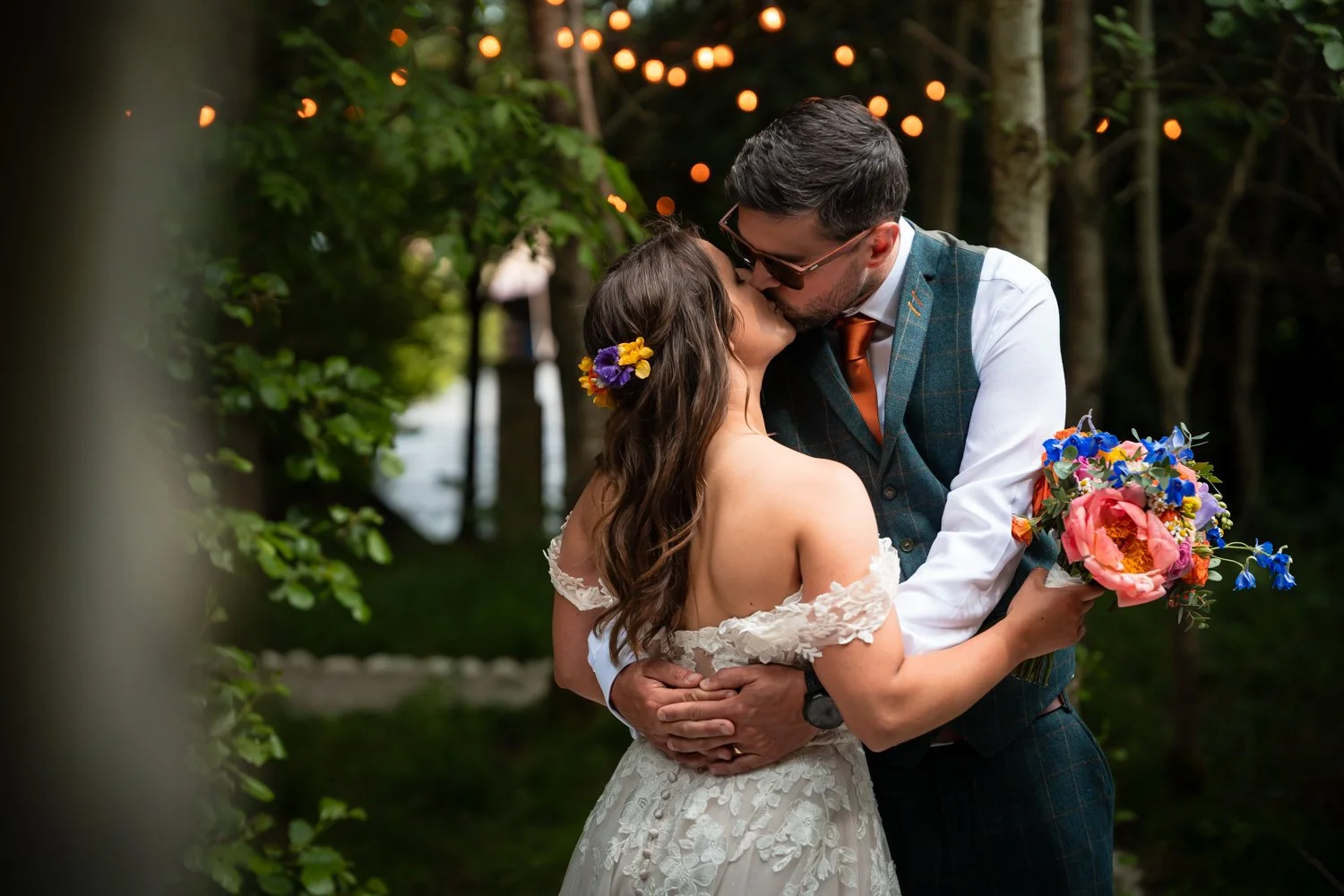 A bride and groom share a kiss outdoors during their wedding, with the bride holding a colorful bouquet and the groom wearing a vest and glasses, surrounded by greenery and string lights.