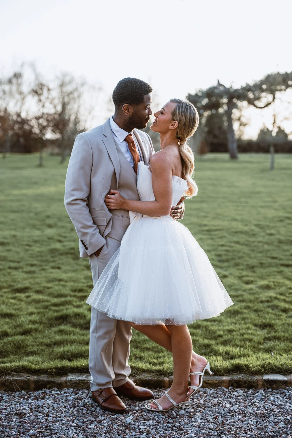 A couple dressed in wedding attire standing close together outdoors on a grassy field during sunset, with trees in the background.