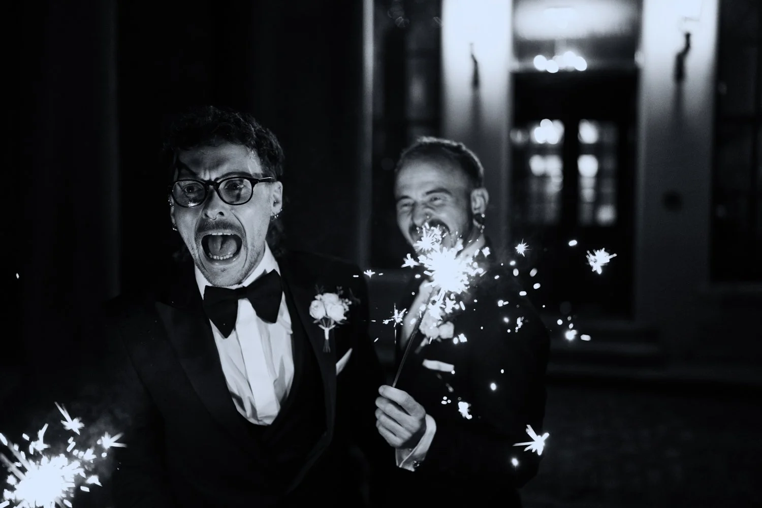 Two men in tuxedos celebrating with sparklers at night, one looking surprised and the other smiling, outside a building.