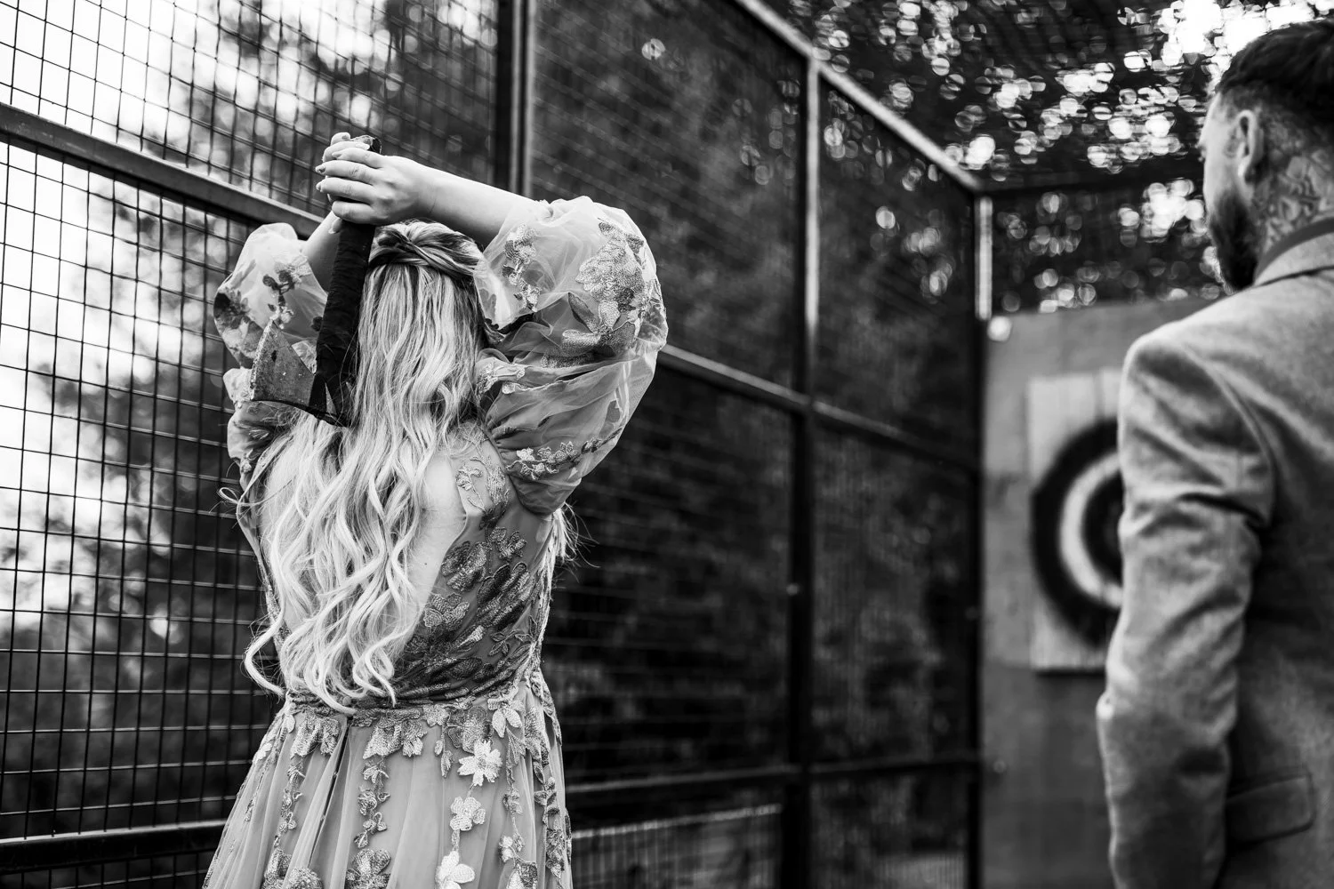 A woman in a floral dress with long wavy hair is standing near a wire fence, with her hands on her head. A man in a suit is standing nearby, facing her, in front of a target.