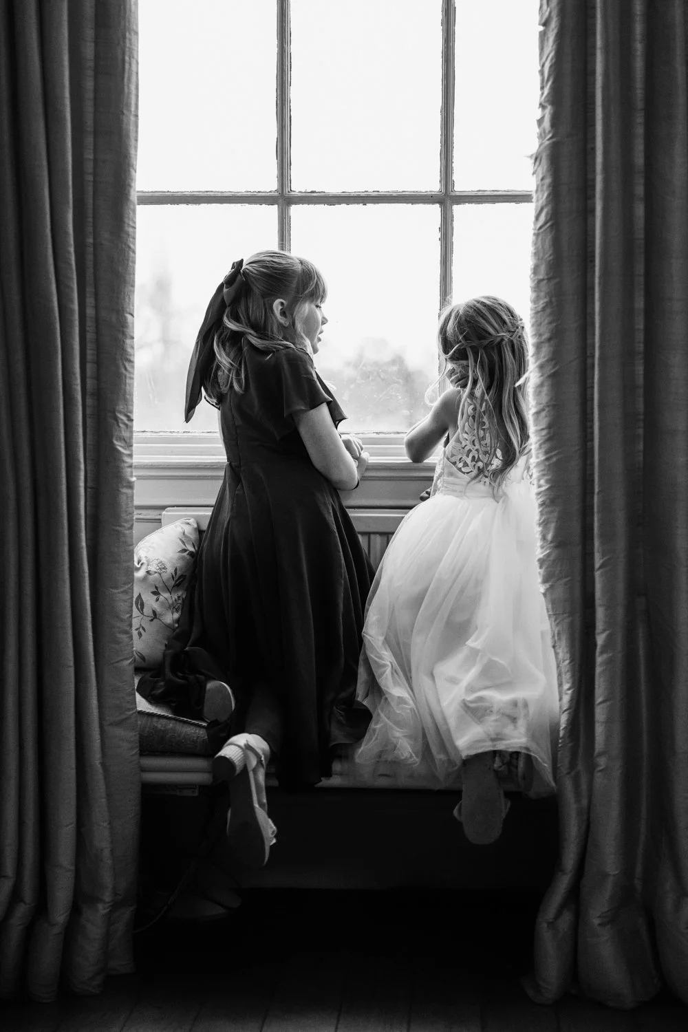 Black and white photo of two young girls standing by a window, one wearing a dark dress and the other in a light, fancy dress, looking outside and talking to each other.