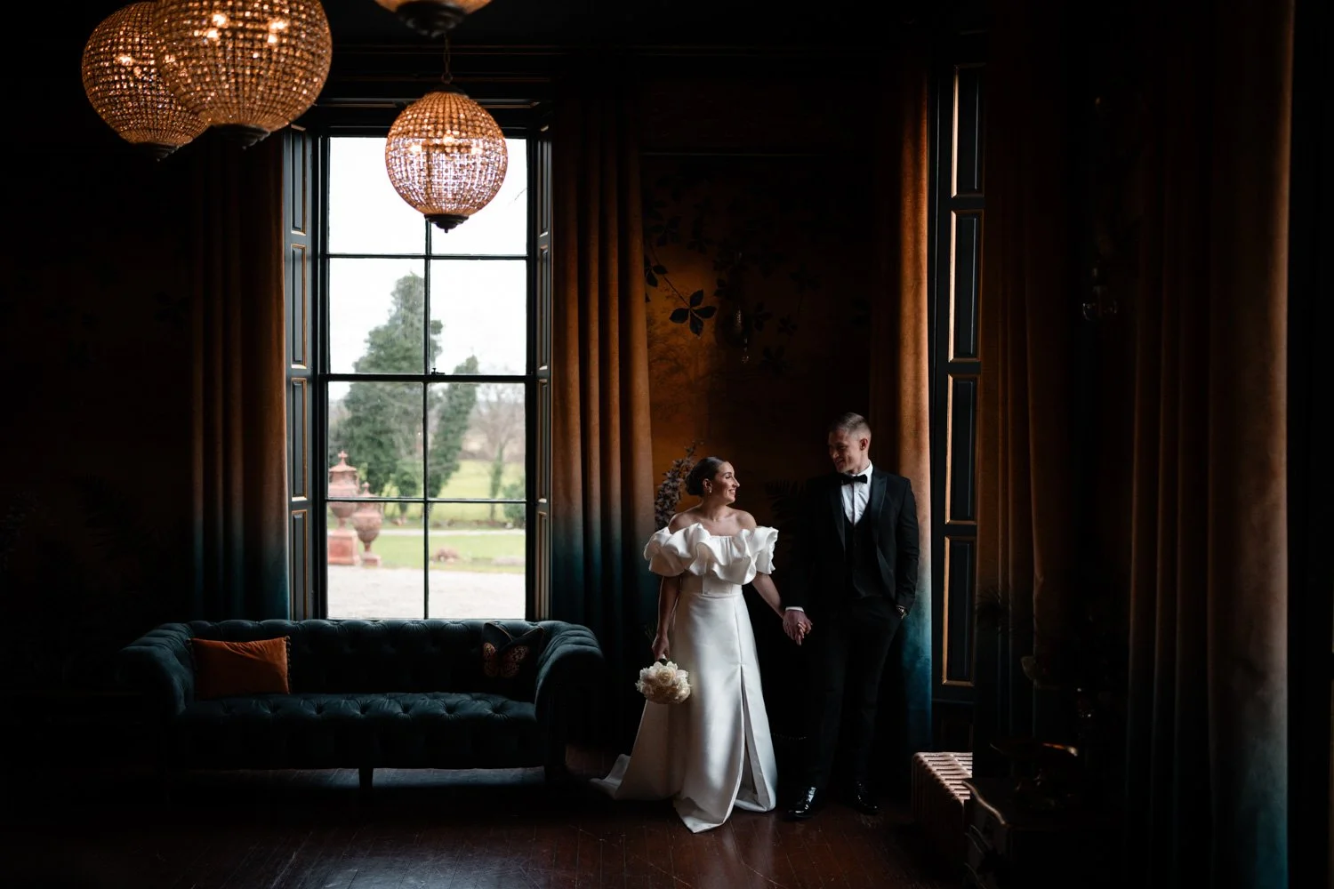A bride and groom holding hands and smiling at each other in a dimly lit room with large windows and elegant chandeliers.