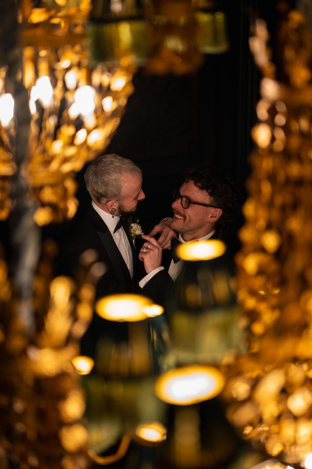 Two men in tuxedos smiling and holding hands at a wedding reception, framed by blurred golden candlelit decorations.